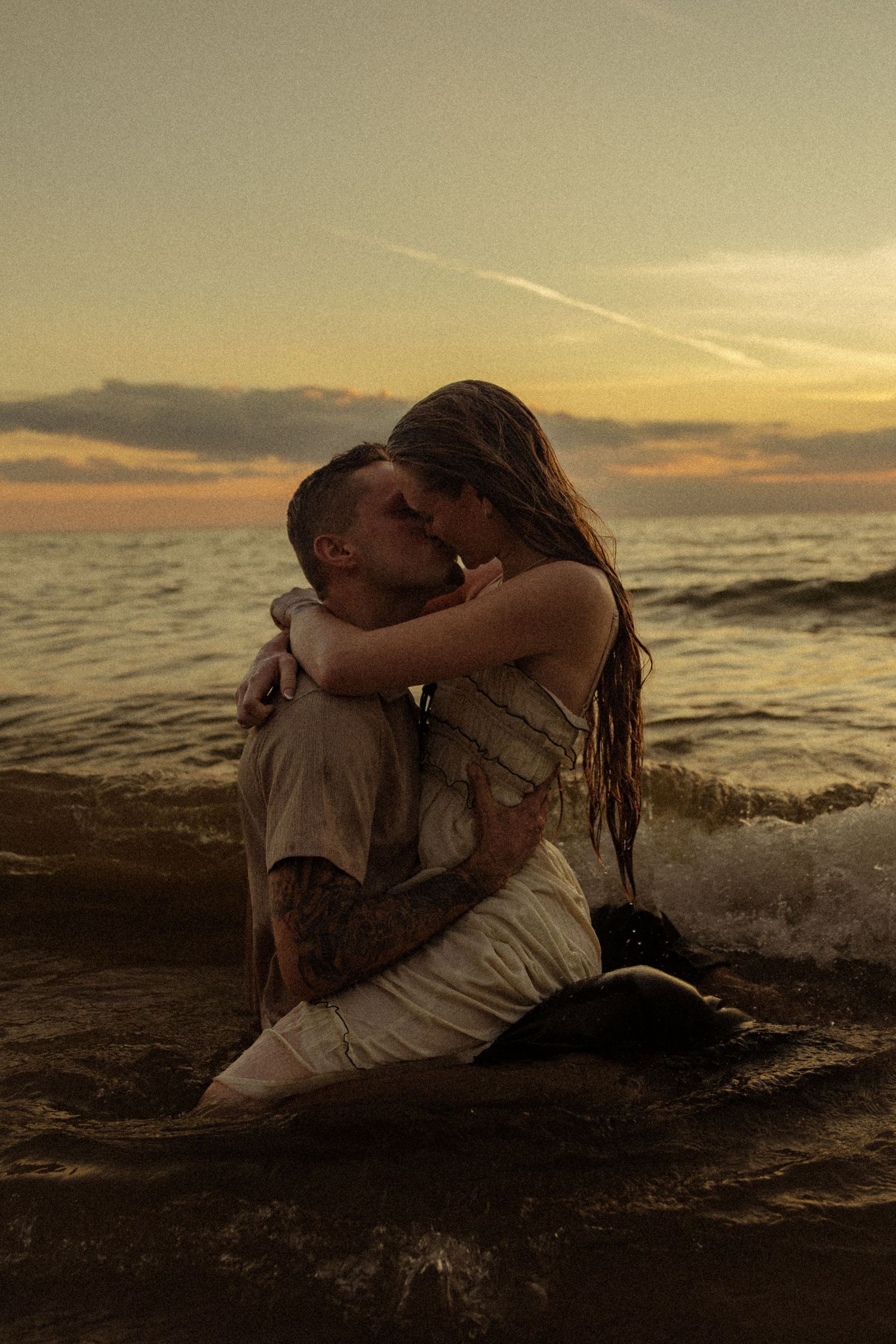 Couple kissing romantically in ocean waves during golden sunset, man with tattoos embracing woman in wet clothing.