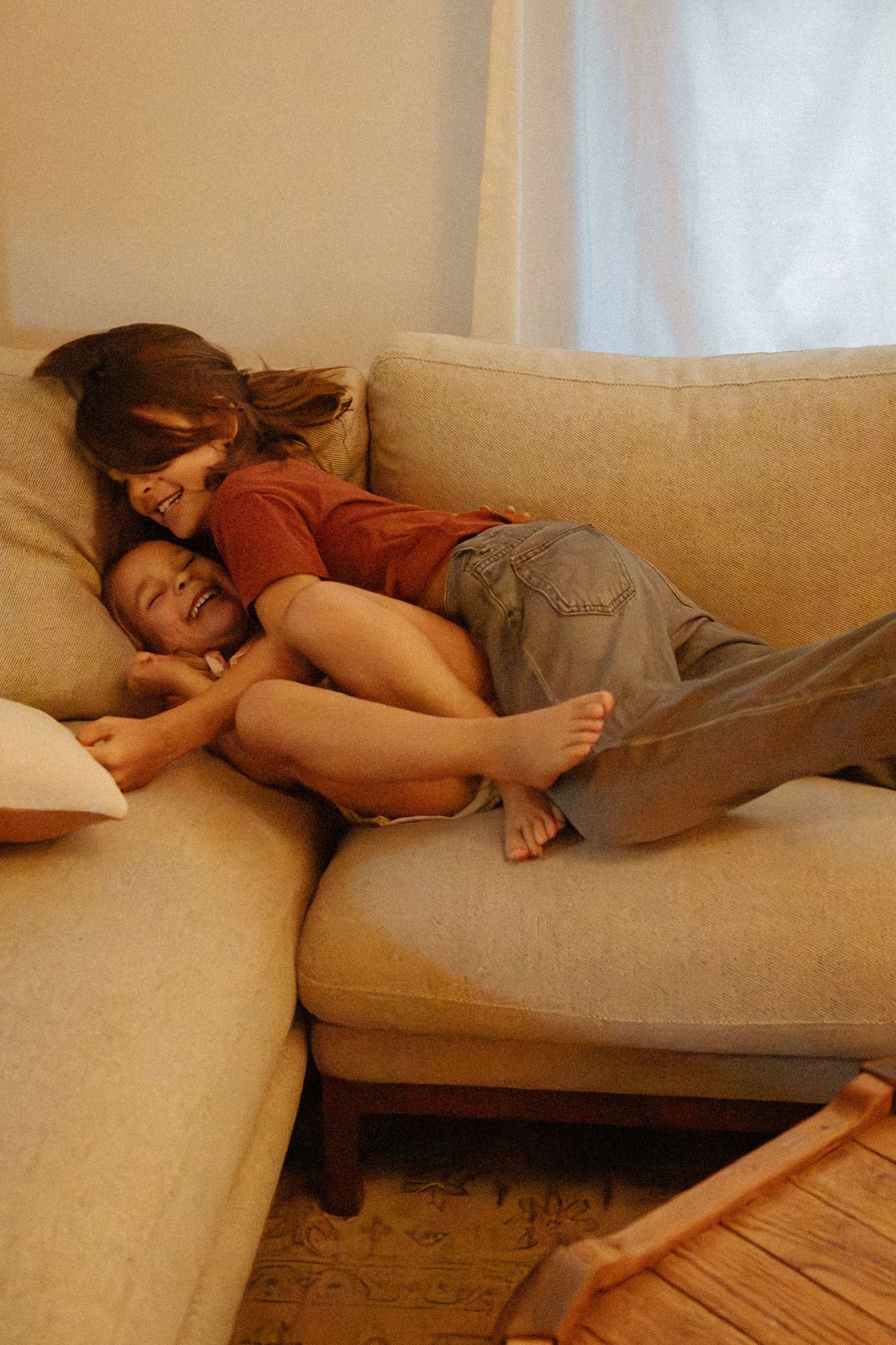 Two children laugh and tumble together on a beige sectional sofa in a softly lit living room near sheer curtains.