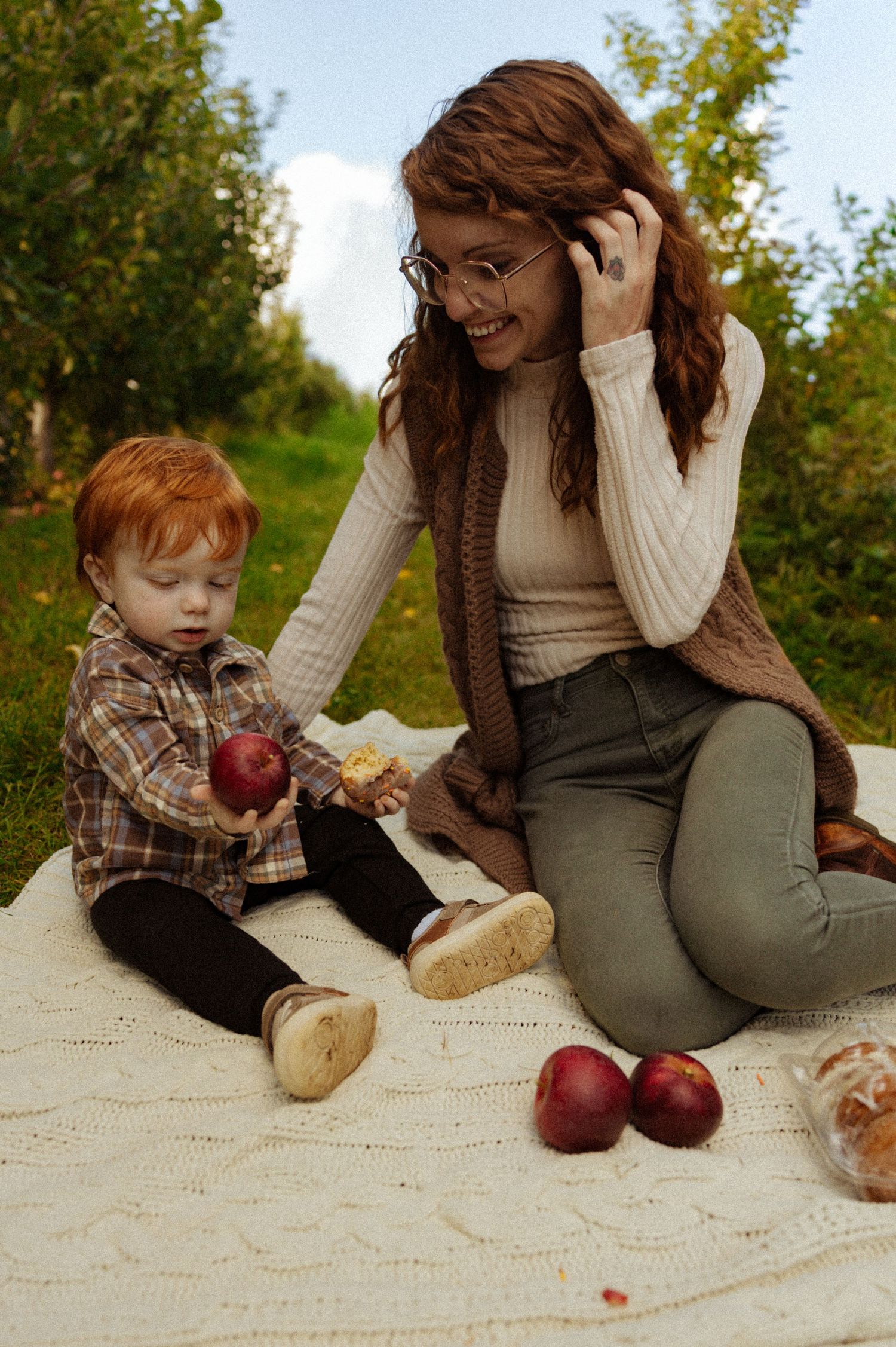 Smiling mother with glasses sitting on blanket with redhead toddler holding apple in orchard.