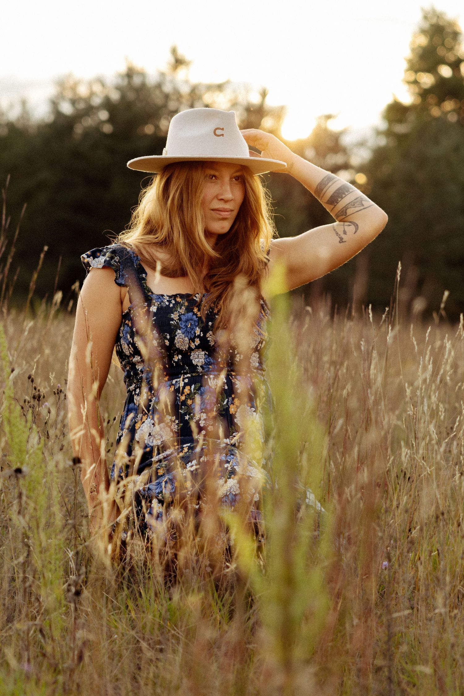 Woman in floral dress and white hat standing in golden tall grass field at sunset with tattoo on arm.