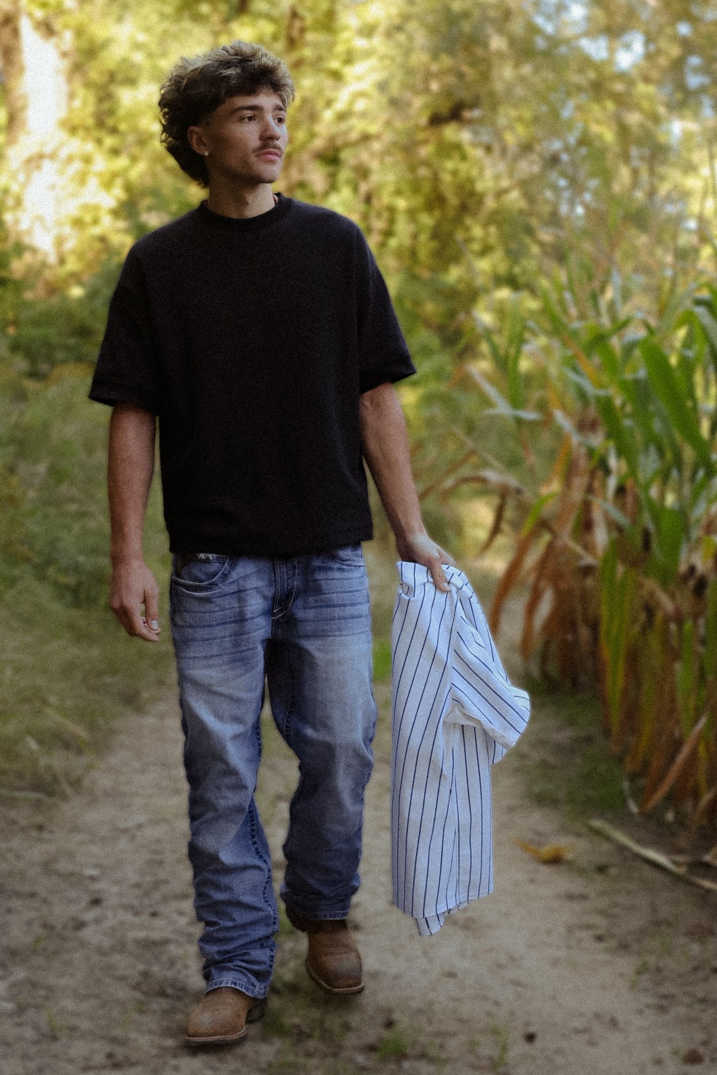 Young man in black t-shirt and jeans walks a dirt path holding a striped shirt, surrounded by lush green trees.