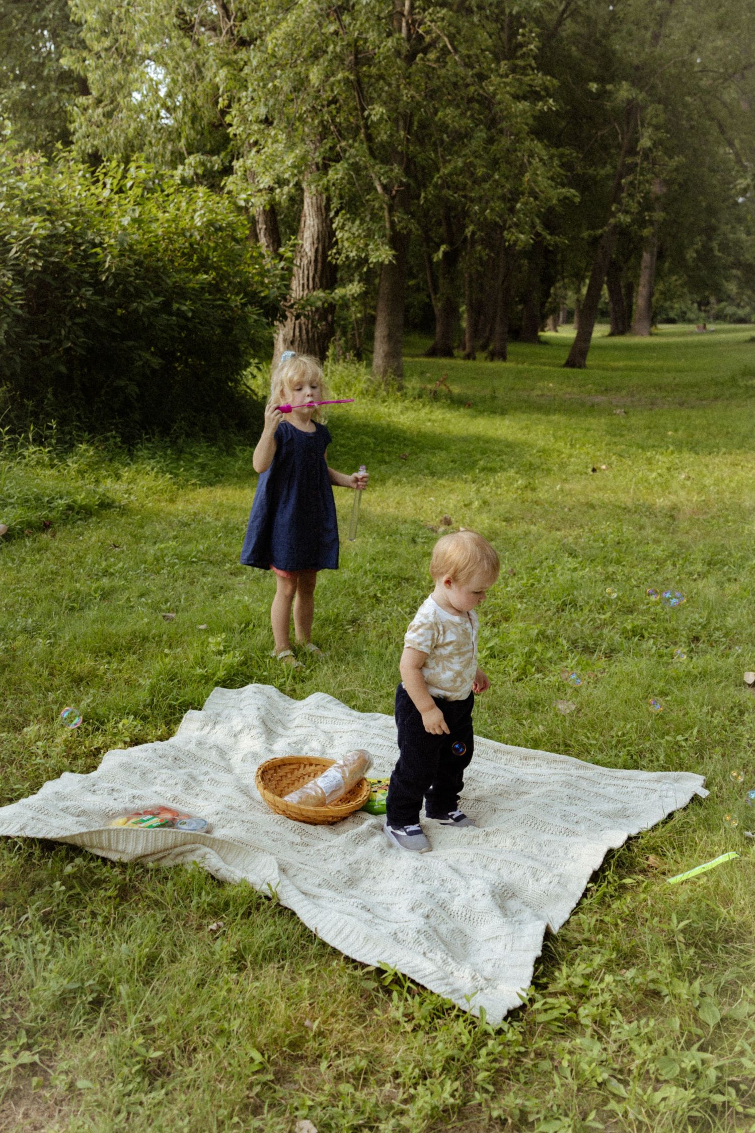 Two young children enjoying a picnic on a blanket in a lush green park, with a wicker basket nearby.