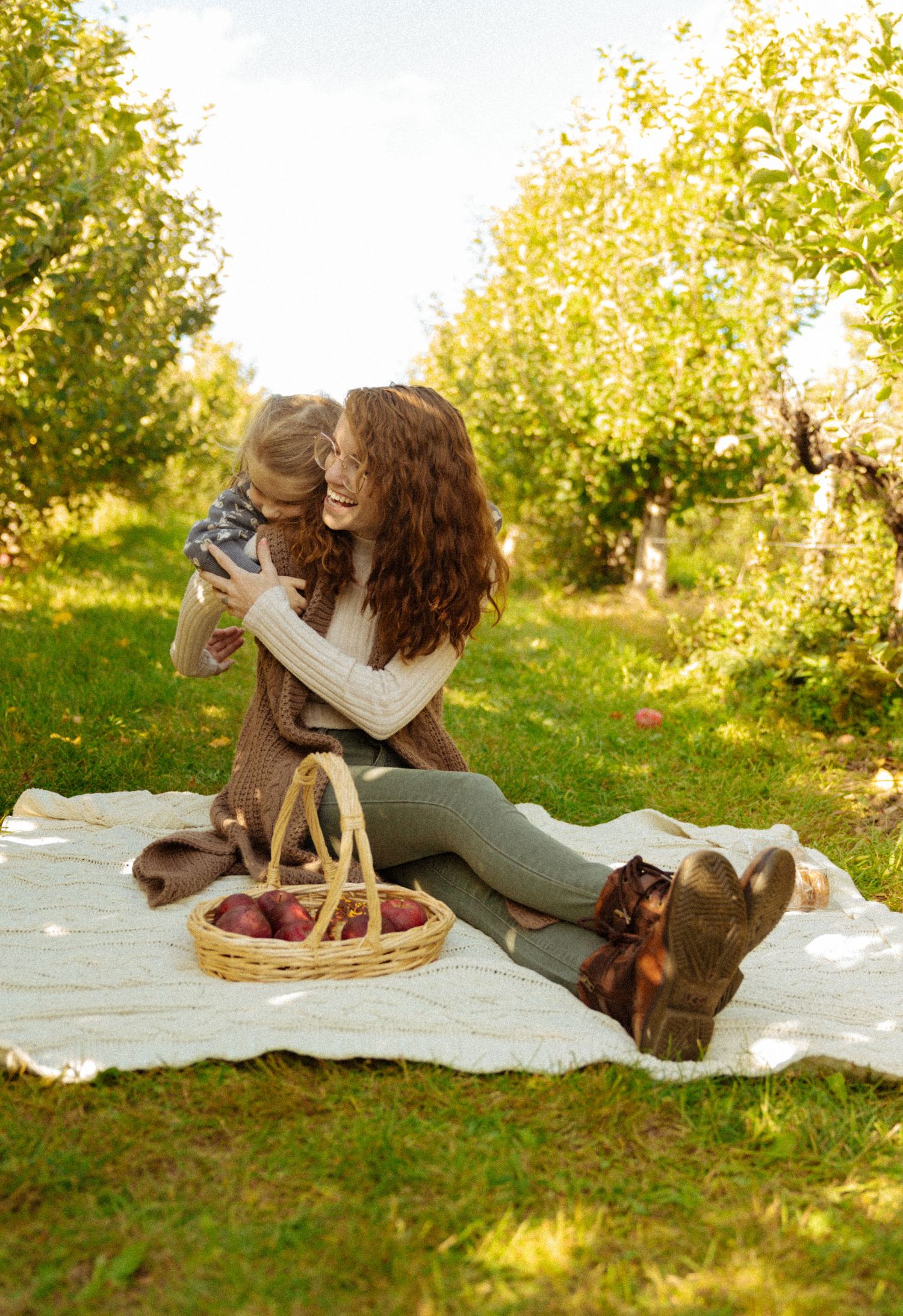 A mother and toddler share a warm hug on a blanket in an apple orchard, with a basket of red apples nearby.