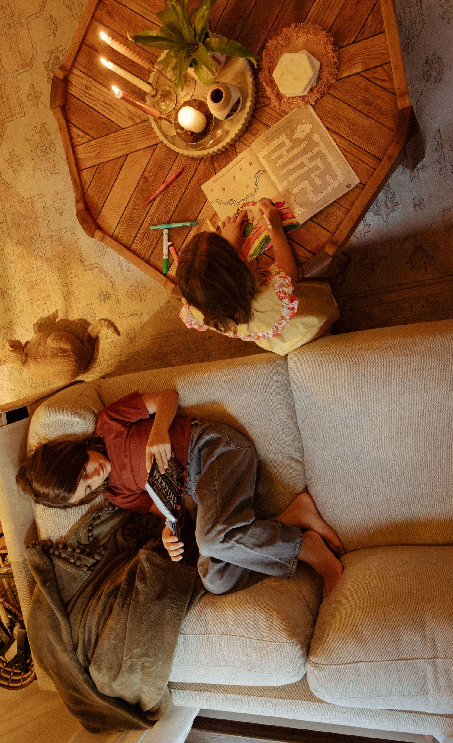 Overhead view of a child reading on a cream sofa while another draws a maze at a wooden coffee table with candles.