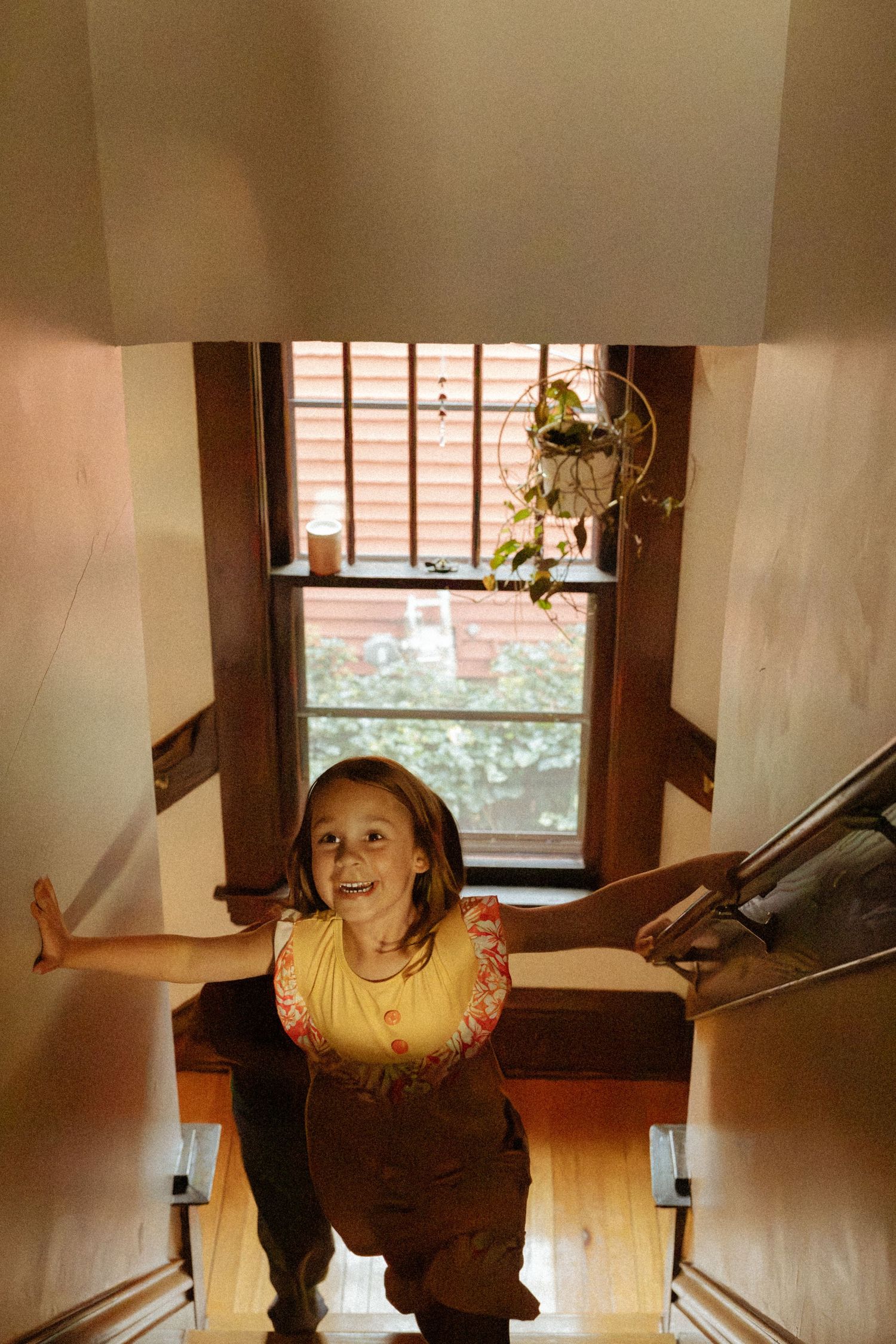 Smiling girl in a yellow floral top climbs a wooden staircase with arms outstretched toward a window with a hanging plant.