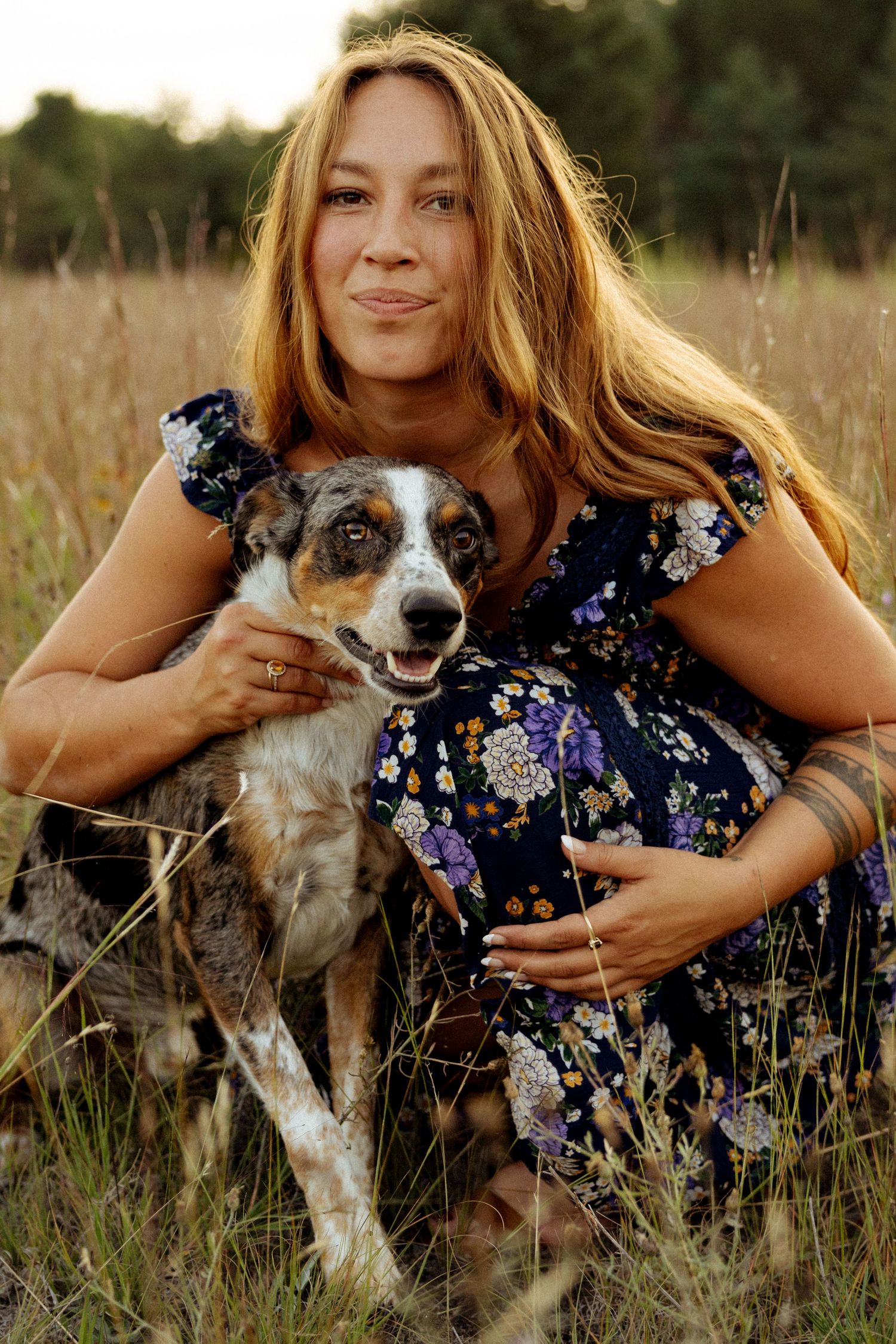 Woman in floral dress hugging her merle Australian Shepherd dog while sitting in a golden grass field.