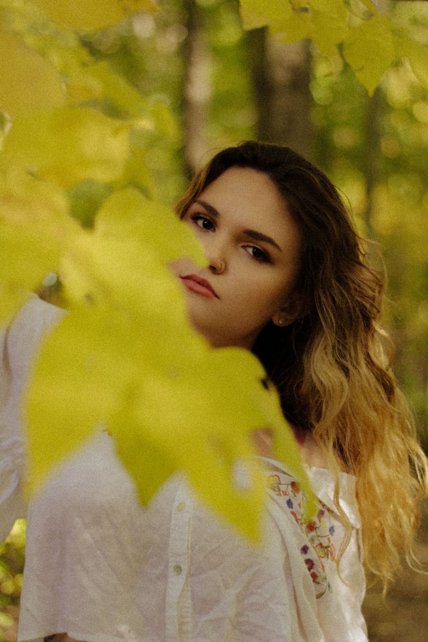 Young woman with wavy hair in white embroidered shirt peers through yellow autumn maple leaves in a forest.