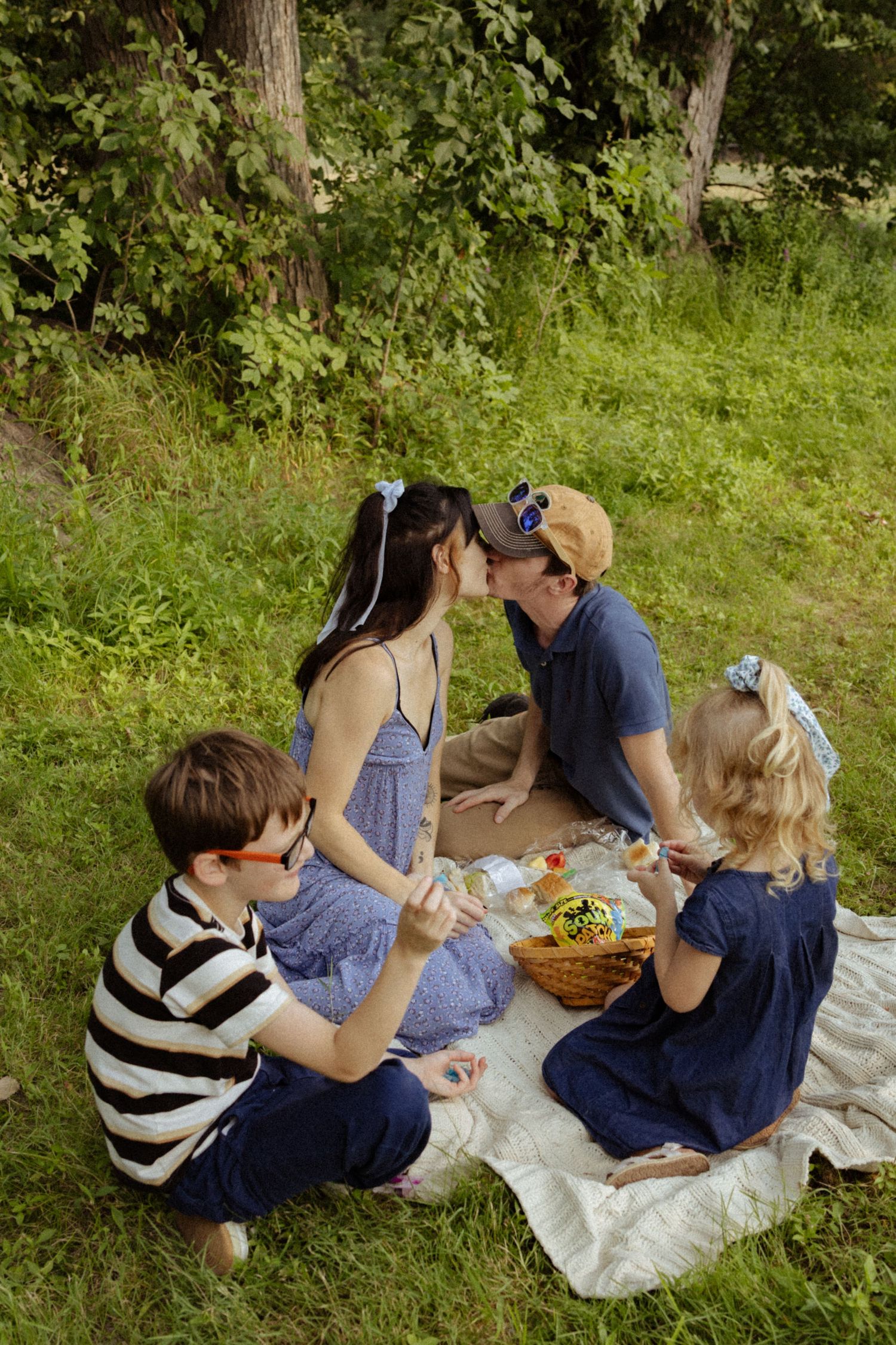 Family of four enjoying a picnic on a blanket in a lush green park, parents sharing a kiss while kids sit nearby.