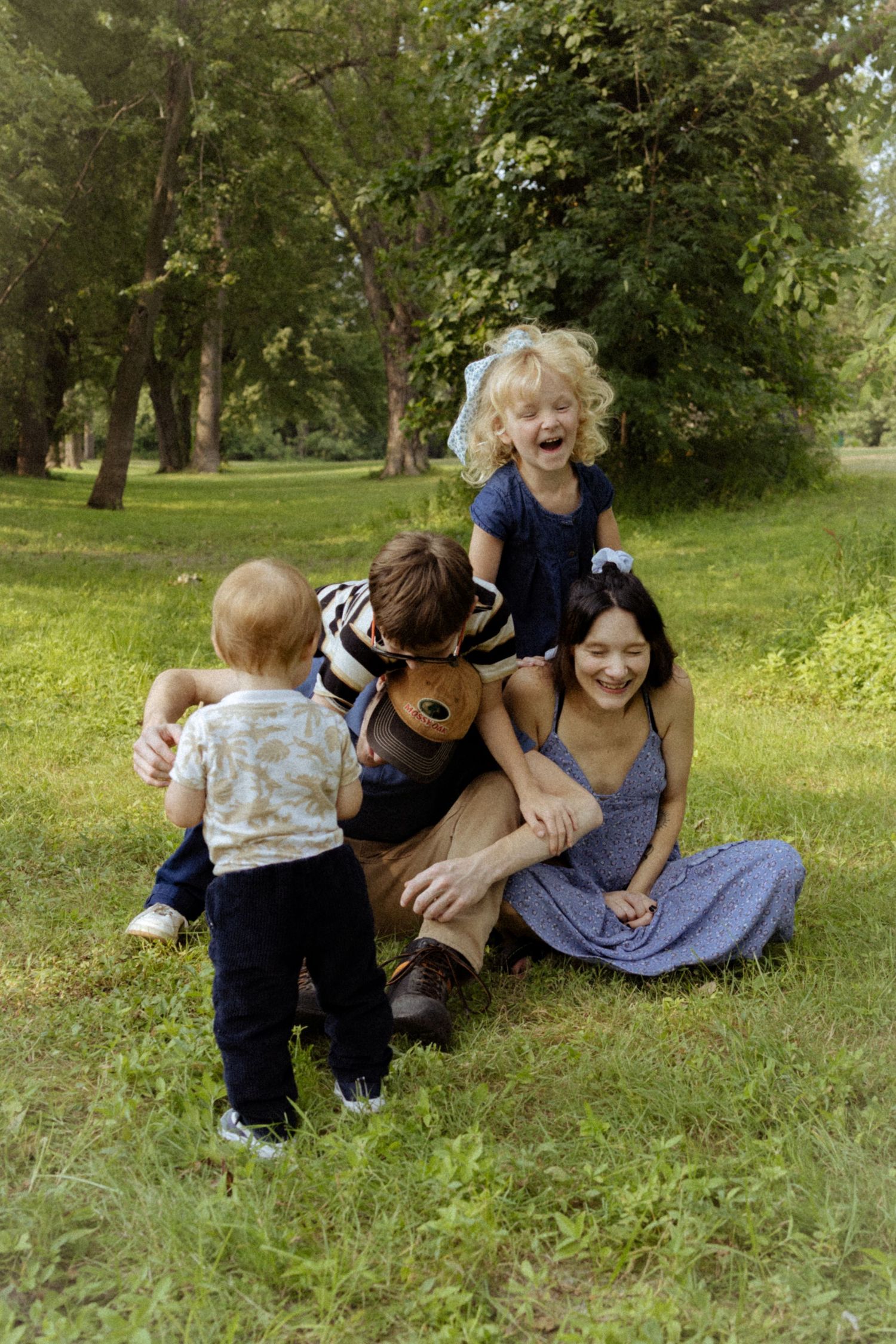 A happy family with two parents and three children playing and laughing together on grass in a park.