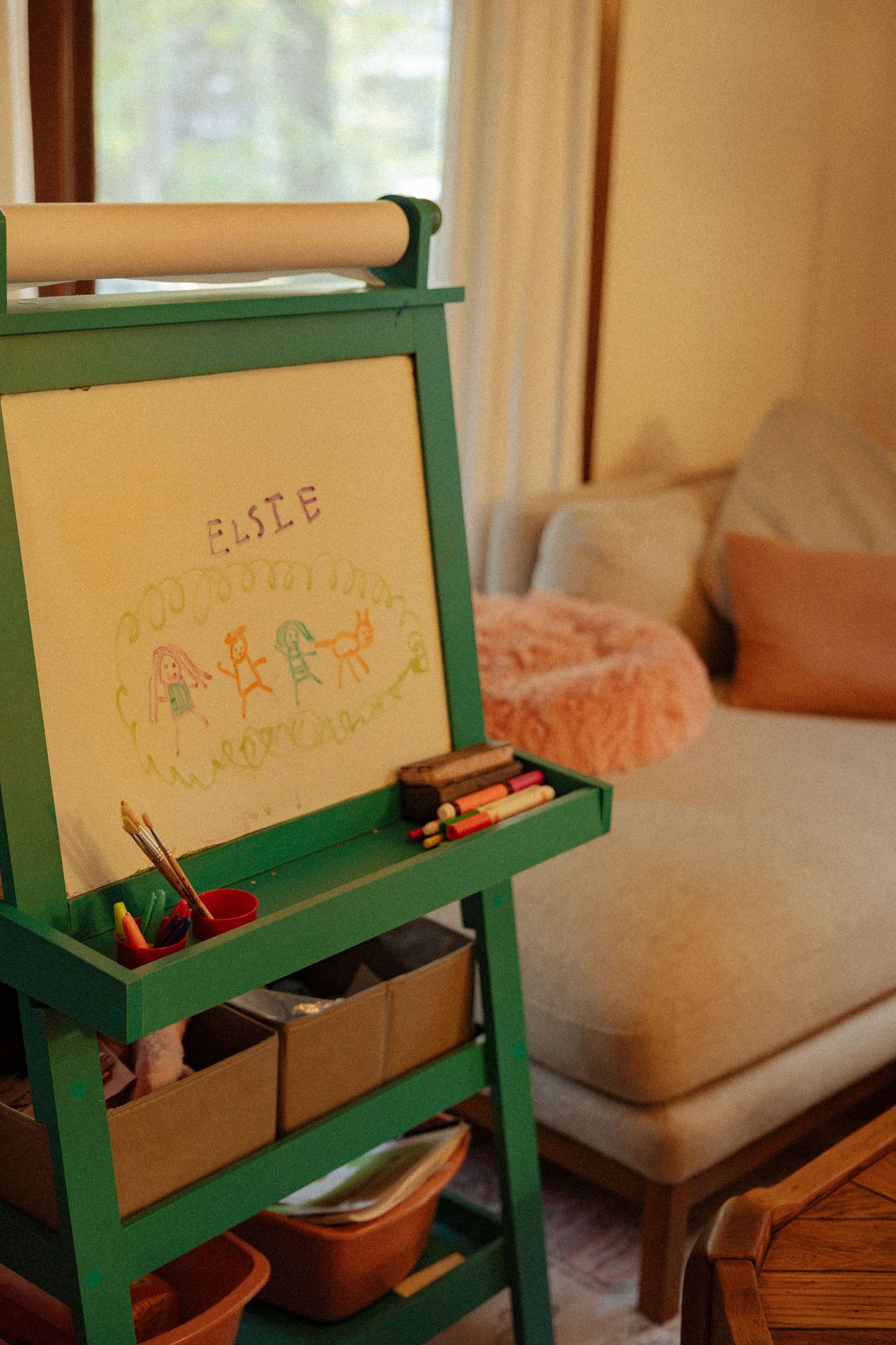 Green wooden children's easel with paper roll and a colorful drawing labeled Elsie, with art supplies in bins beside a cozy sofa.