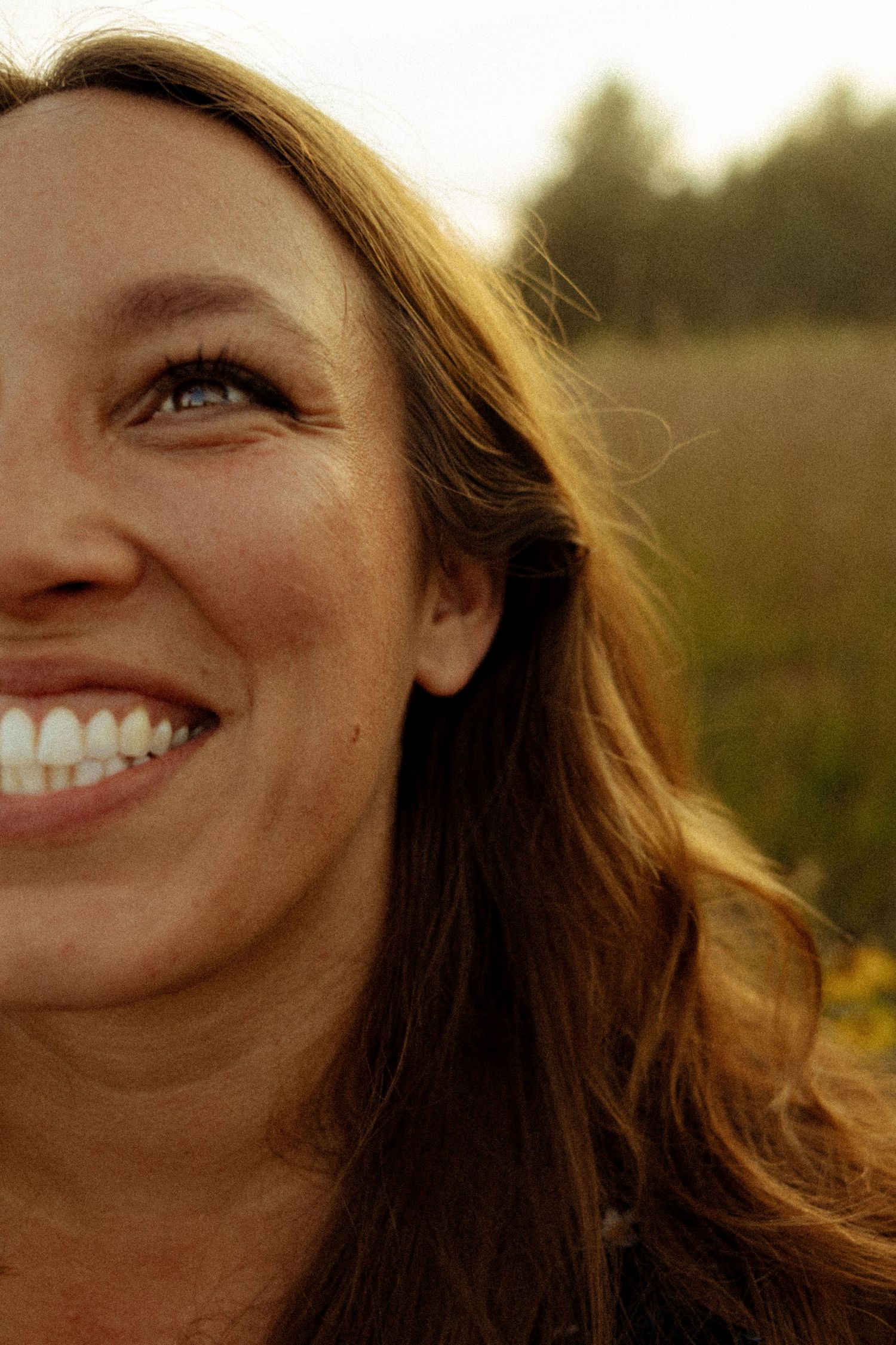 A smiling woman with long brown hair and blue eyes enjoys a sunny outdoor moment in a natural field setting.
