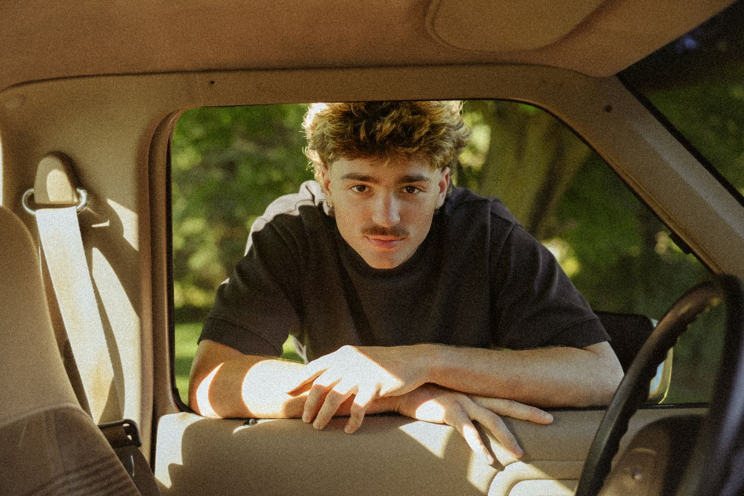 Young man with mustache leaning on car seat interior, casual black tee, surrounded by green trees in natural light.