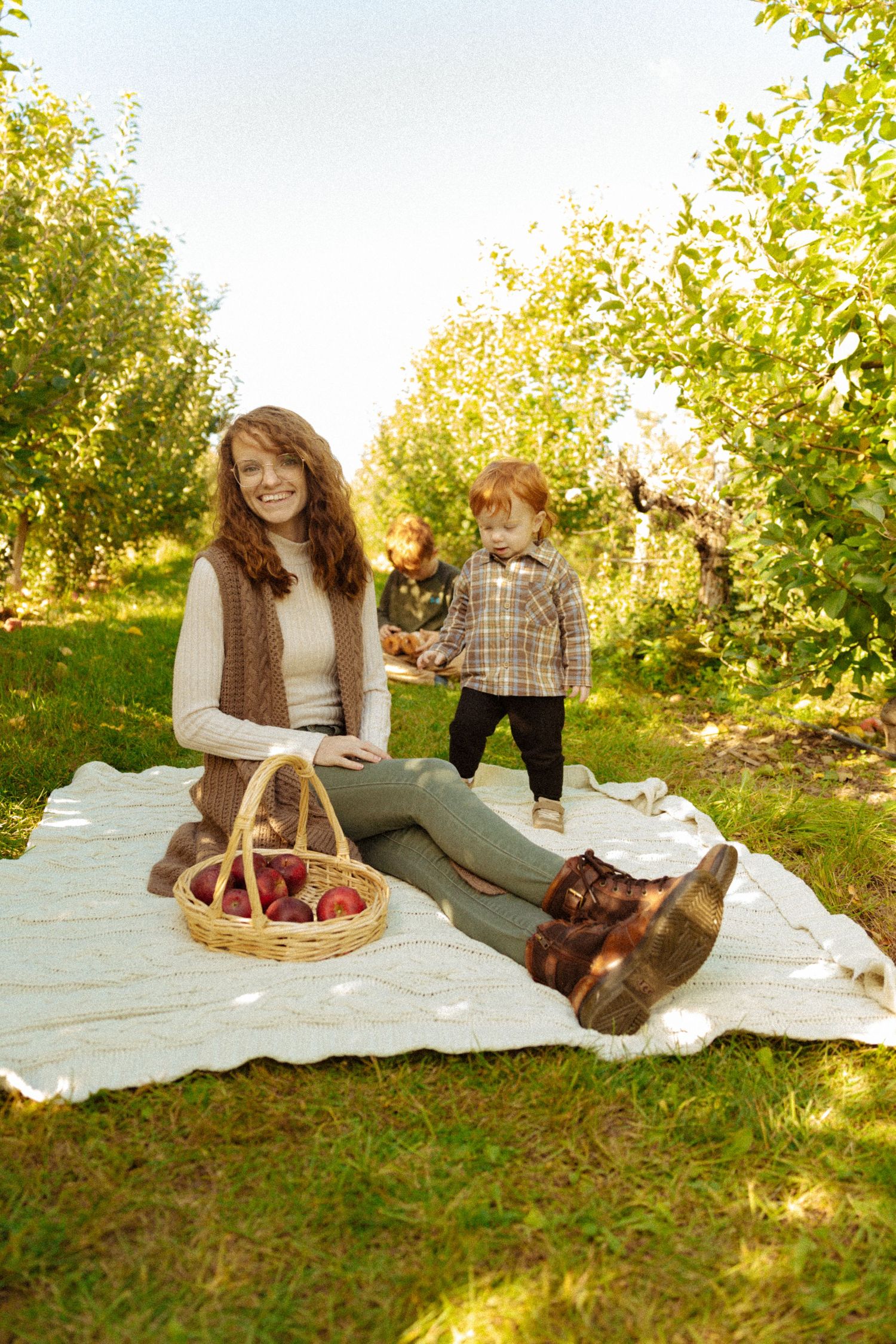 Woman sitting on blanket in apple orchard with basket of red apples and toddler standing nearby on sunny day.