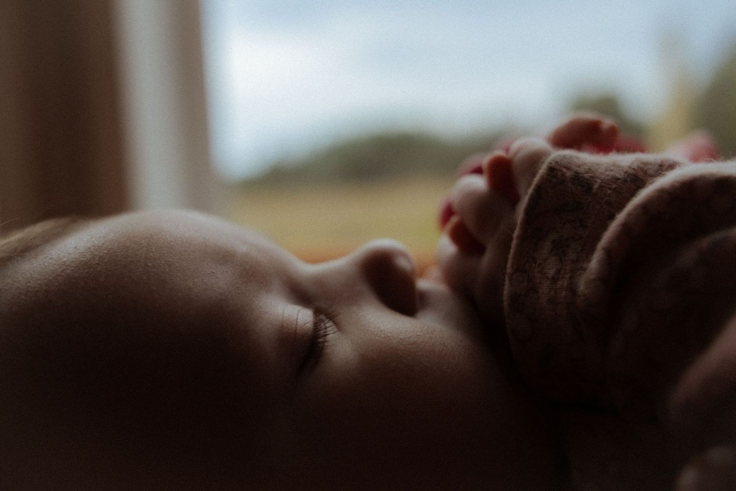 Close-up of a sleeping newborn baby with soft skin, resting peacefully near a window in natural light.