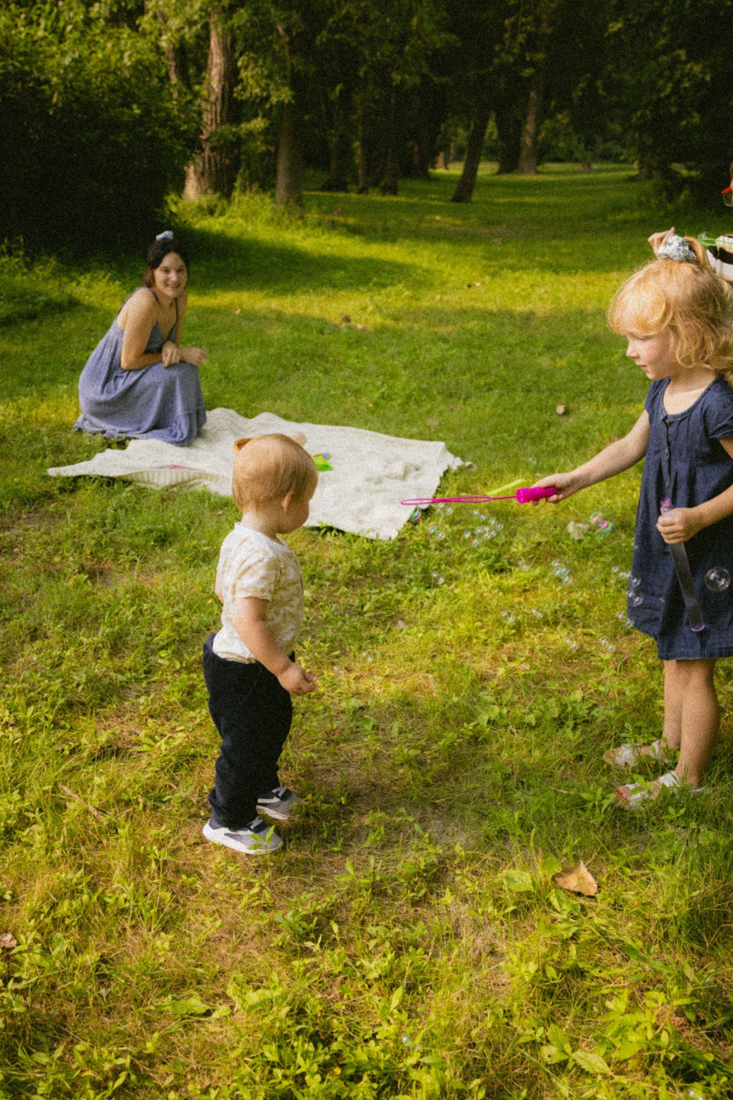 Two children play with bubbles on grass in a park while a woman sits on a blanket in the background.