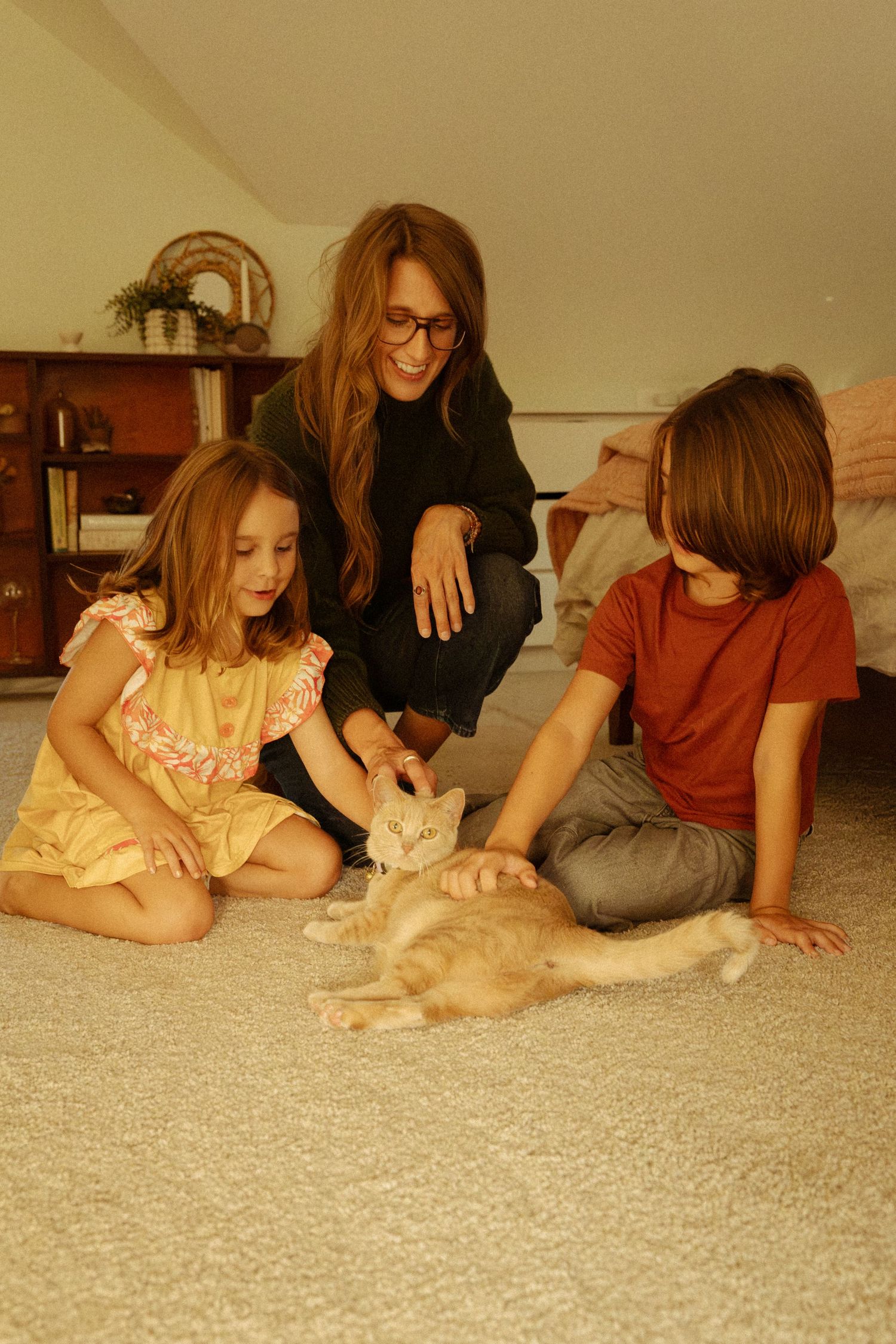 A smiling woman with glasses and two children sit on a soft carpet petting an orange cat in a cozy attic-style bedroom.