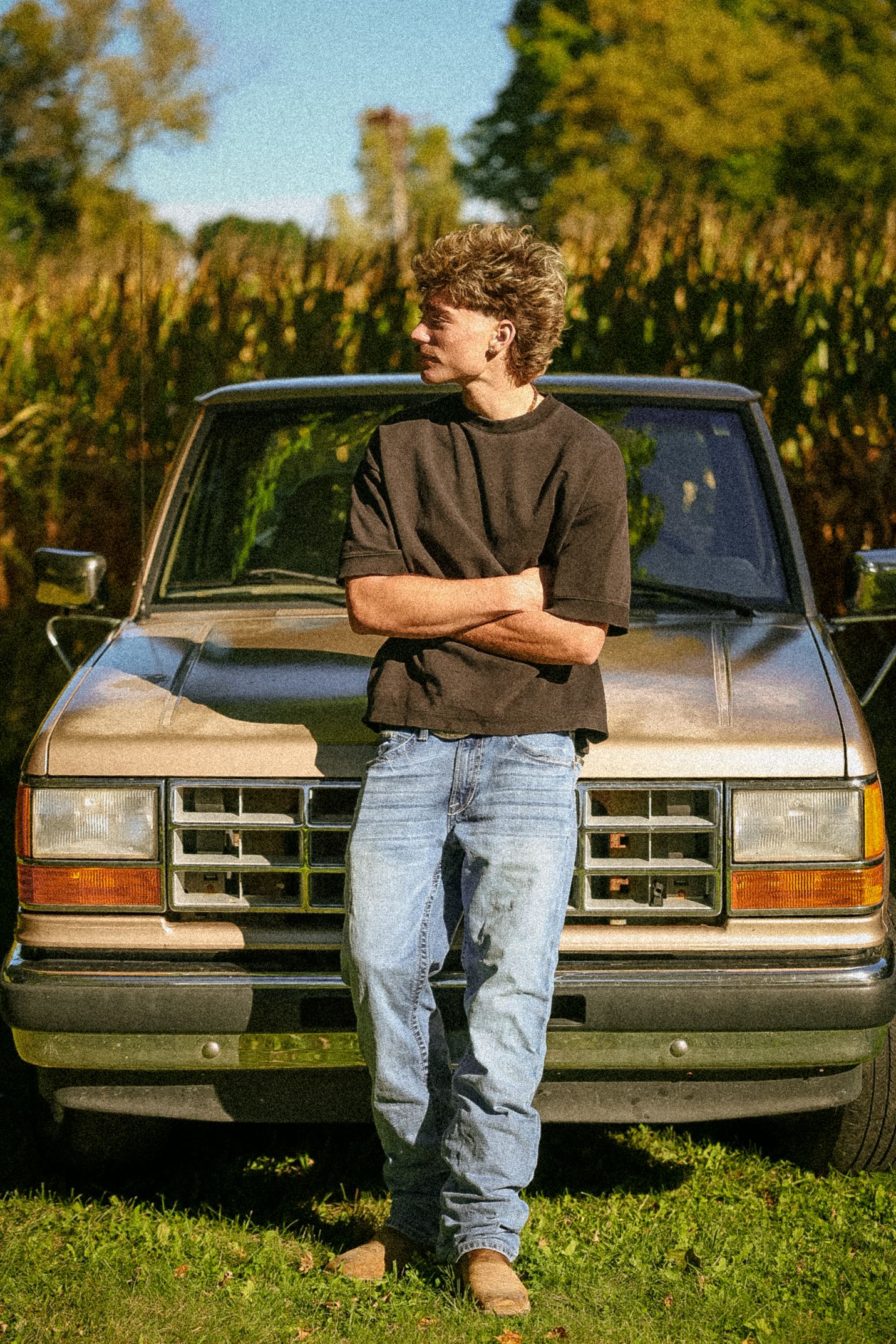 Young man with curly hair in black tee and jeans leans against vintage truck near cornfield on sunny day.