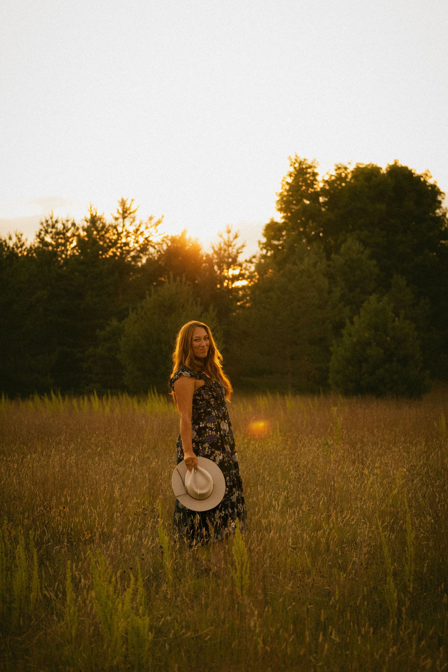 Woman in floral maxi dress holding hat standing in golden sunset meadow with trees in background.