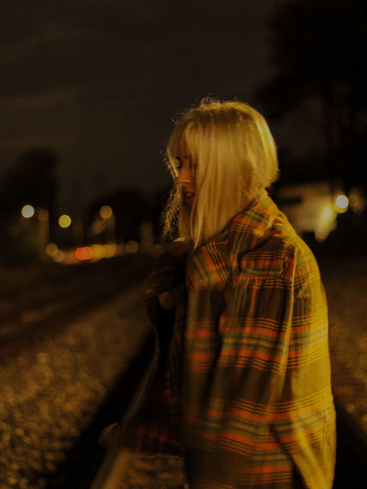 Blonde woman in plaid jacket standing on railroad tracks at night with bokeh city lights in background.