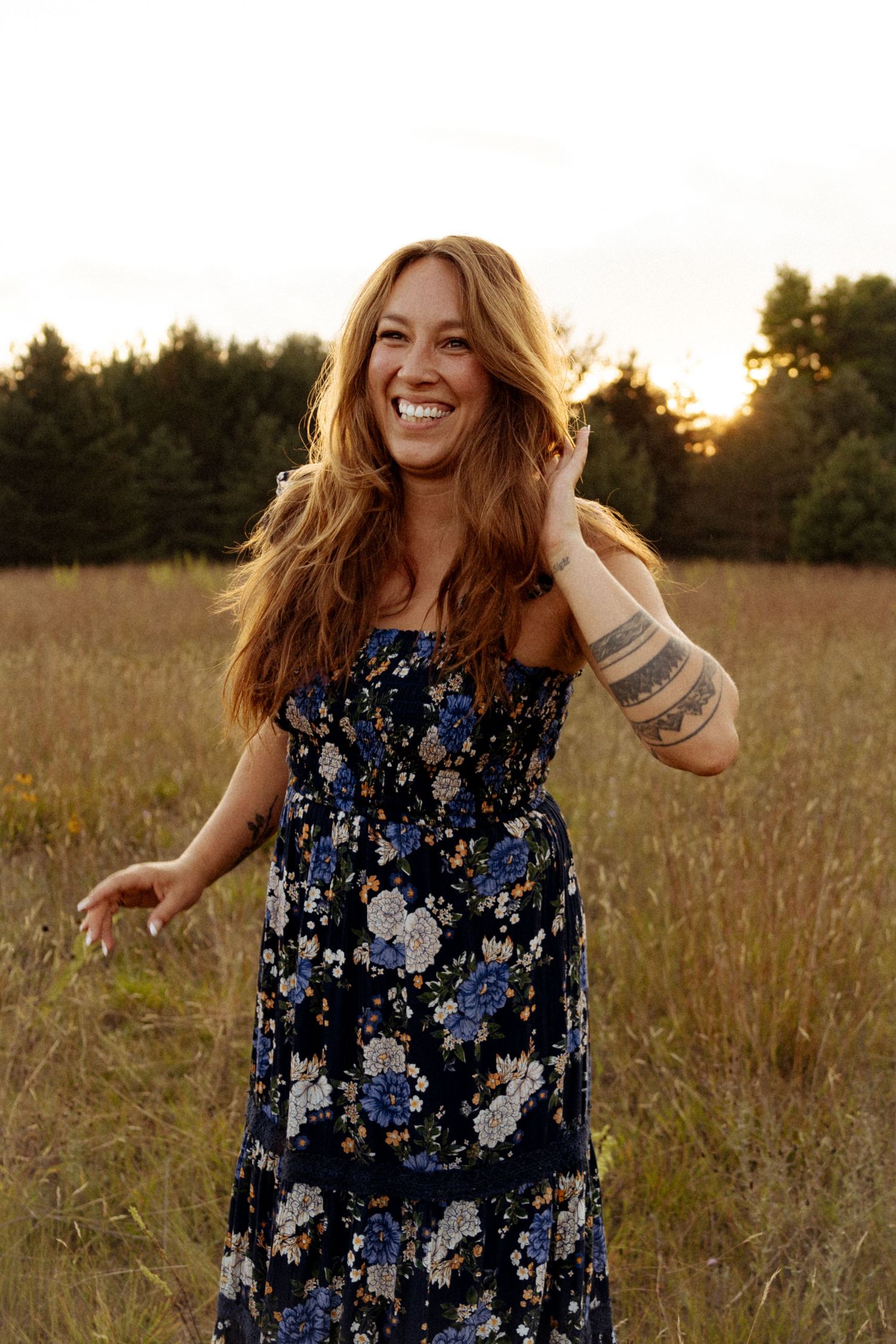 Smiling woman in floral maxi dress with arm tattoo laughing in golden field at sunset.