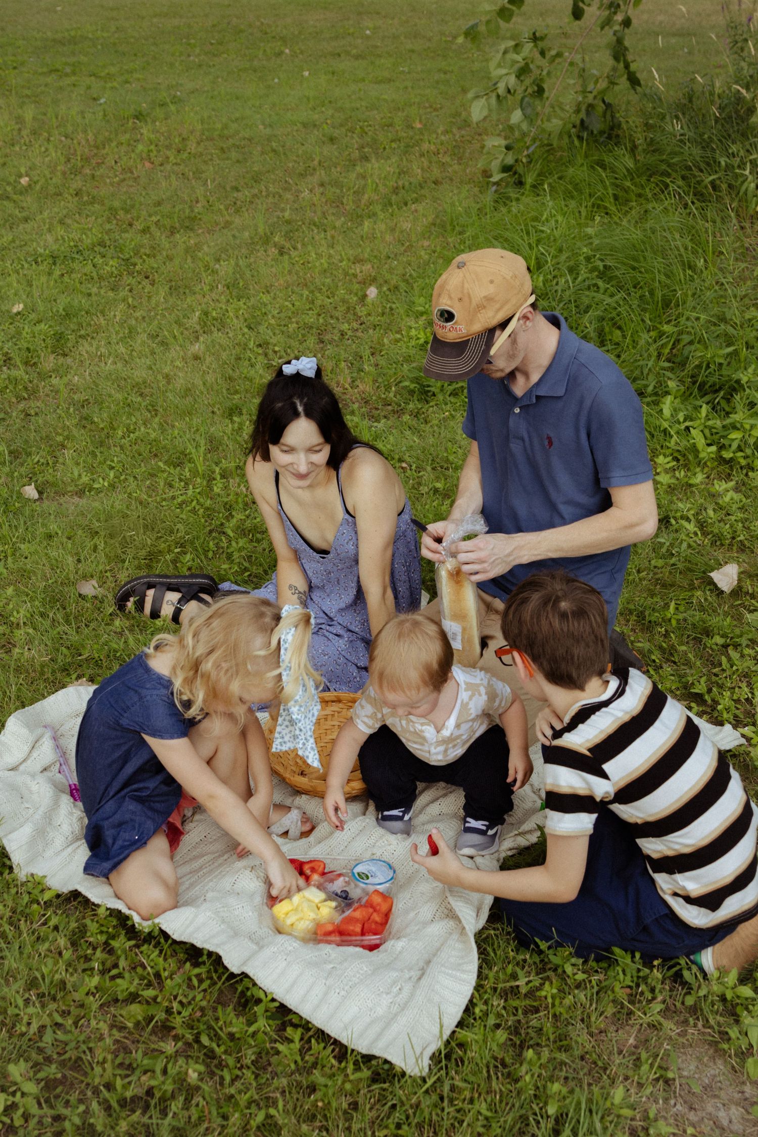 Family of five sharing a picnic on a blanket in a green park with a basket of fresh fruit.
