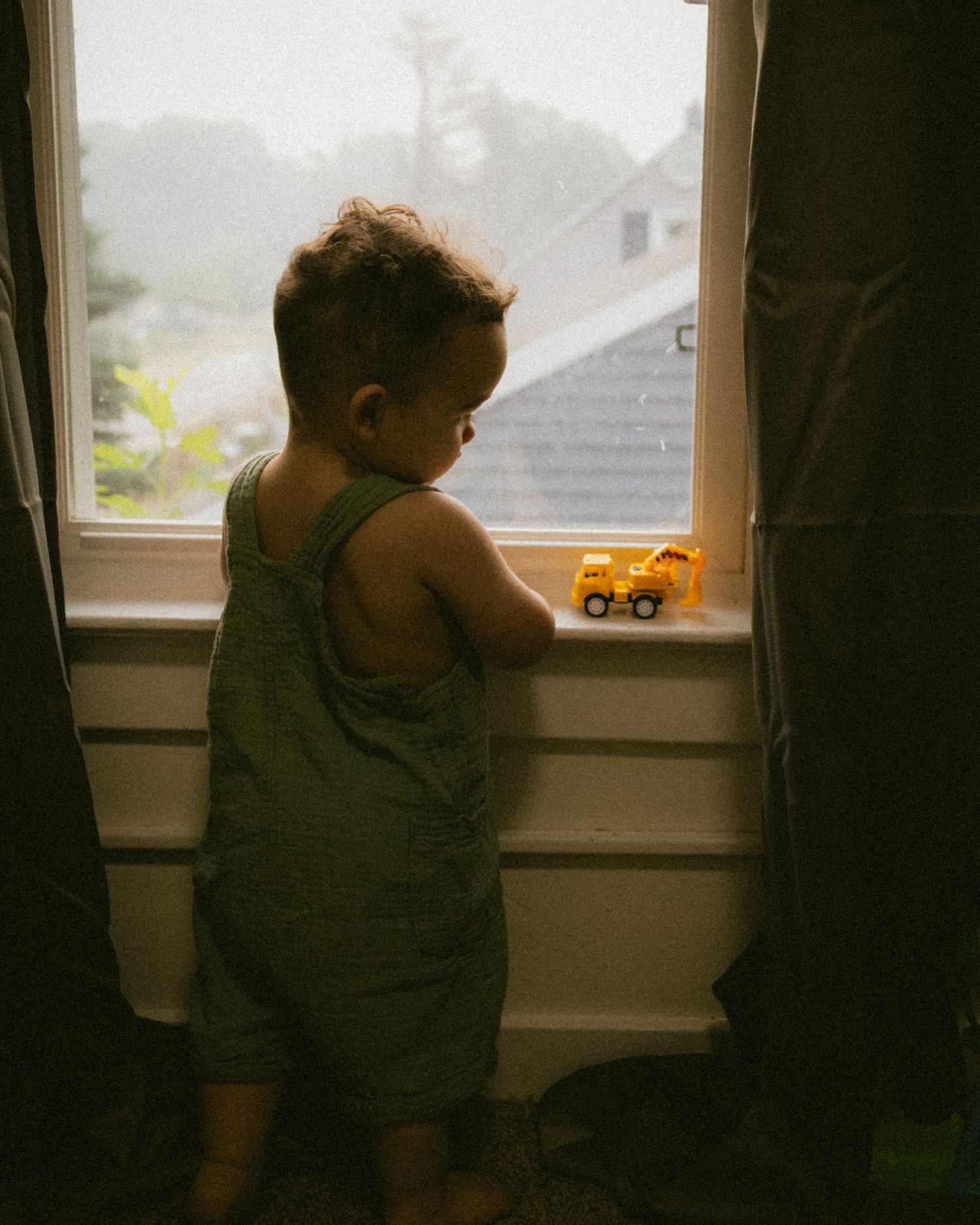 Toddler in green overalls playing with a yellow toy truck on a windowsill while looking outside on a rainy day.