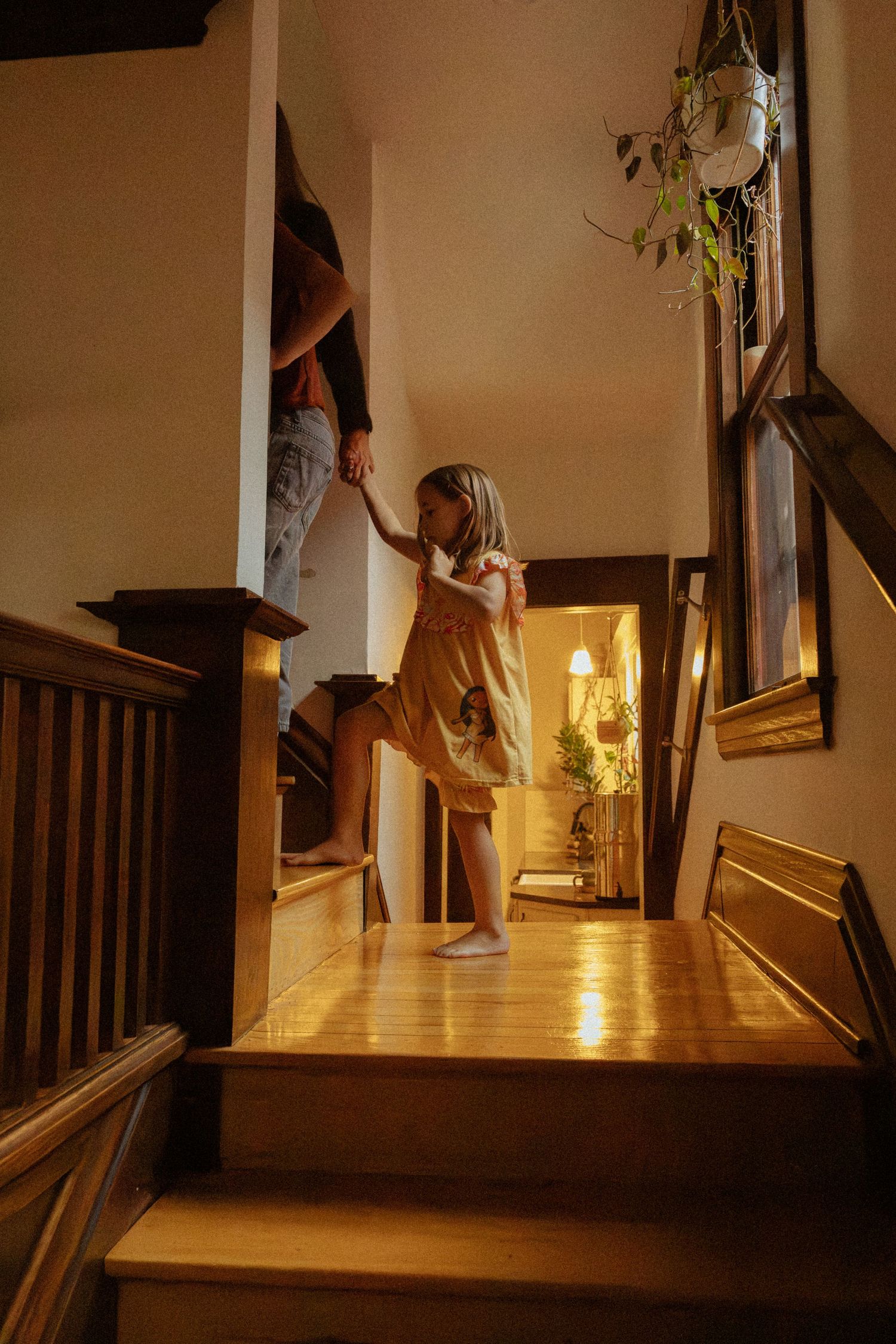 A young girl in a yellow dress stands barefoot on a wooden staircase landing, holding an adult's hand in a warm, softly lit home.