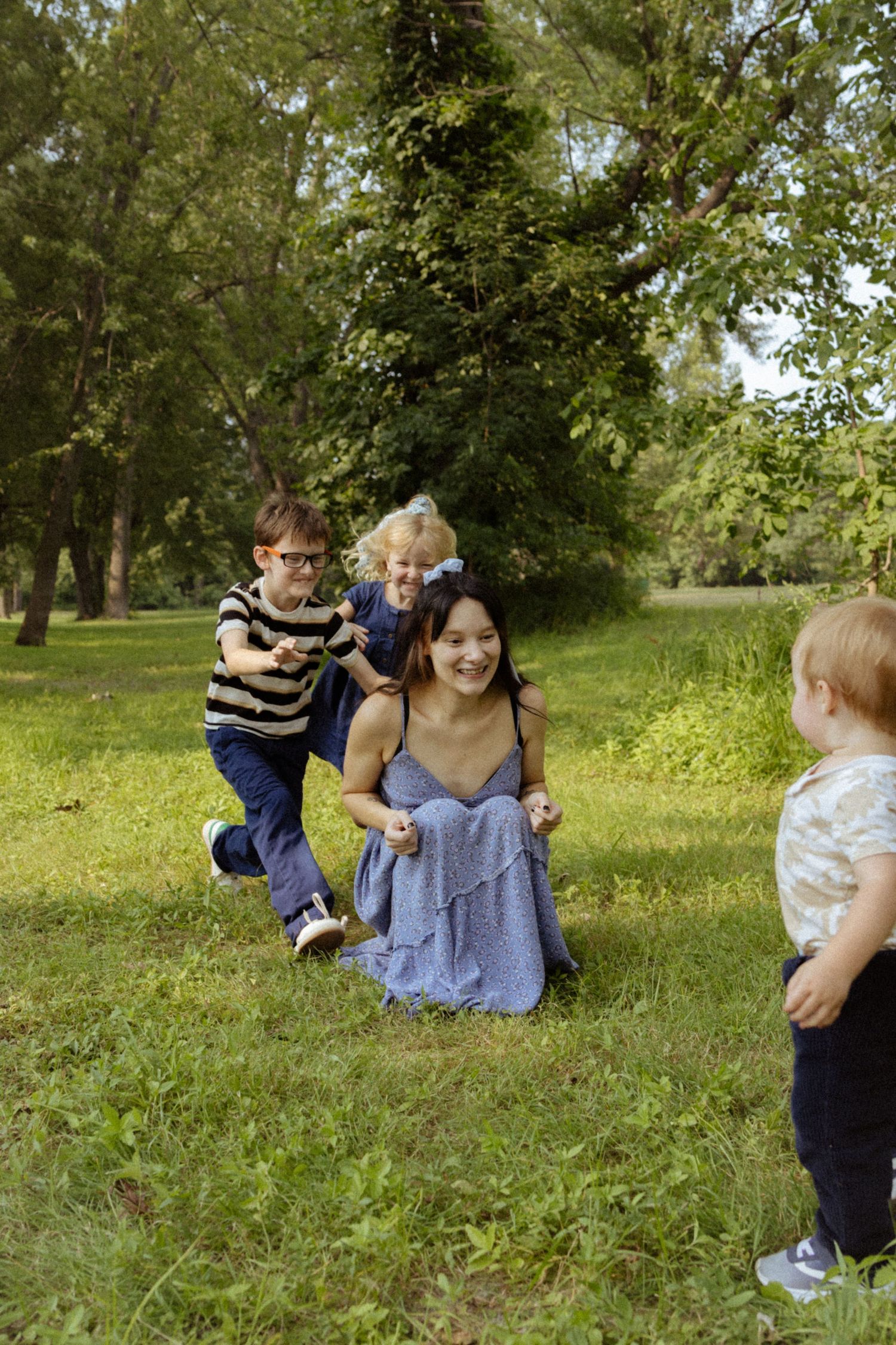 A smiling woman sits on grass in a park while three young children play around her on a sunny day.