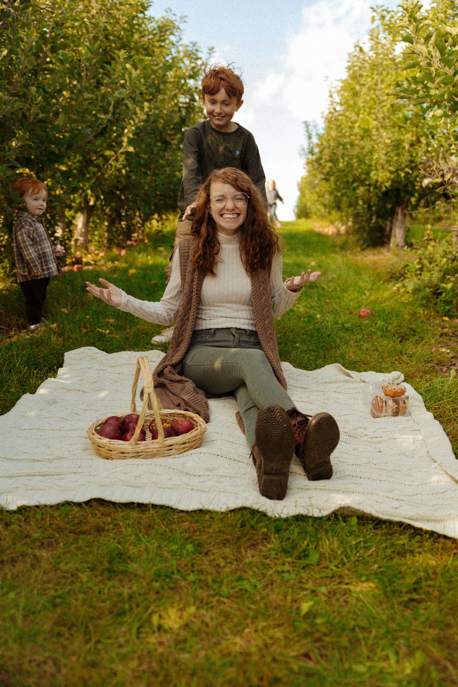 Woman sitting on blanket in apple orchard smiling with two children playing behind her near a basket of apples.