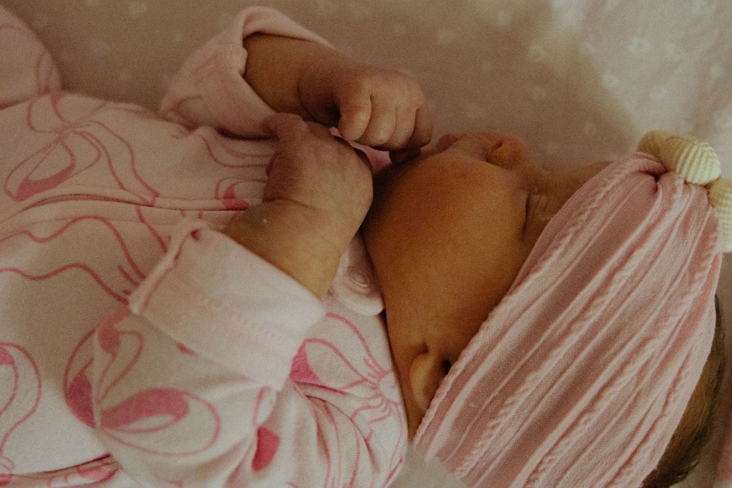 Newborn baby sleeping peacefully in pink floral outfit, tiny hands visible, resting in a white bassinet.