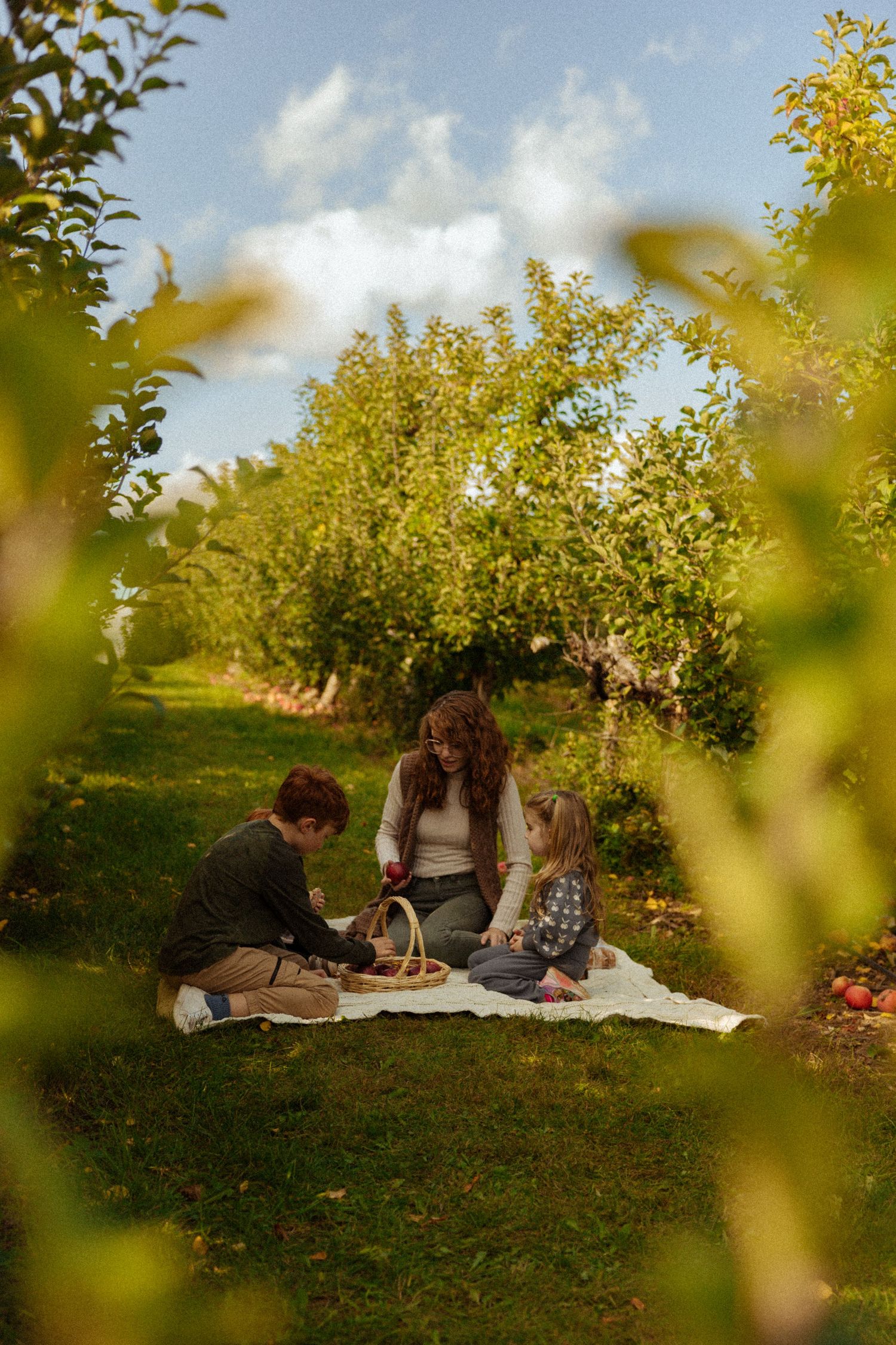 Family enjoying a picnic in a lush apple orchard on a sunny day with a wicker basket.