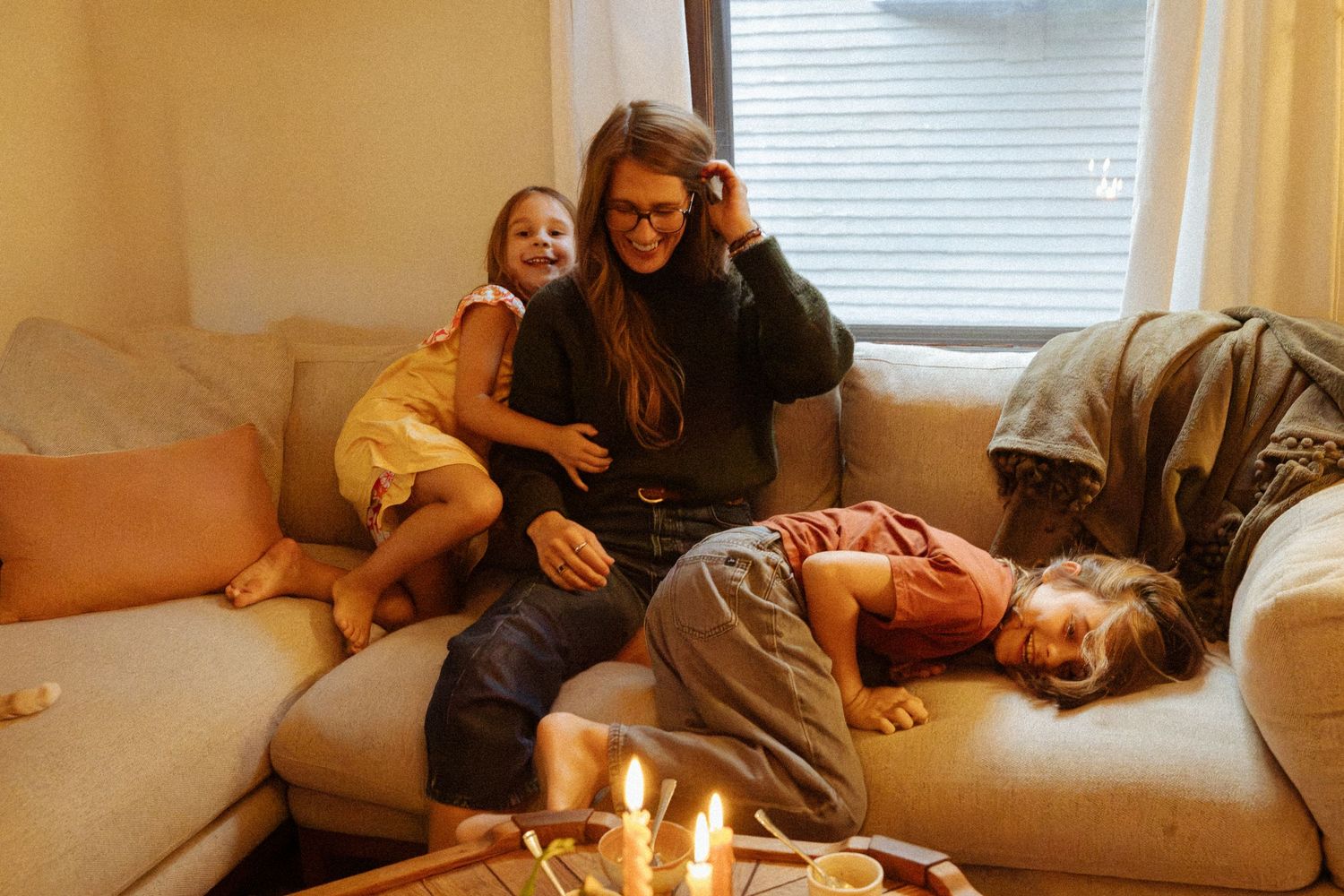 A woman sits on a cozy sofa smiling as two children snuggle and play beside her, with lit candles glowing on a table in the foreground.