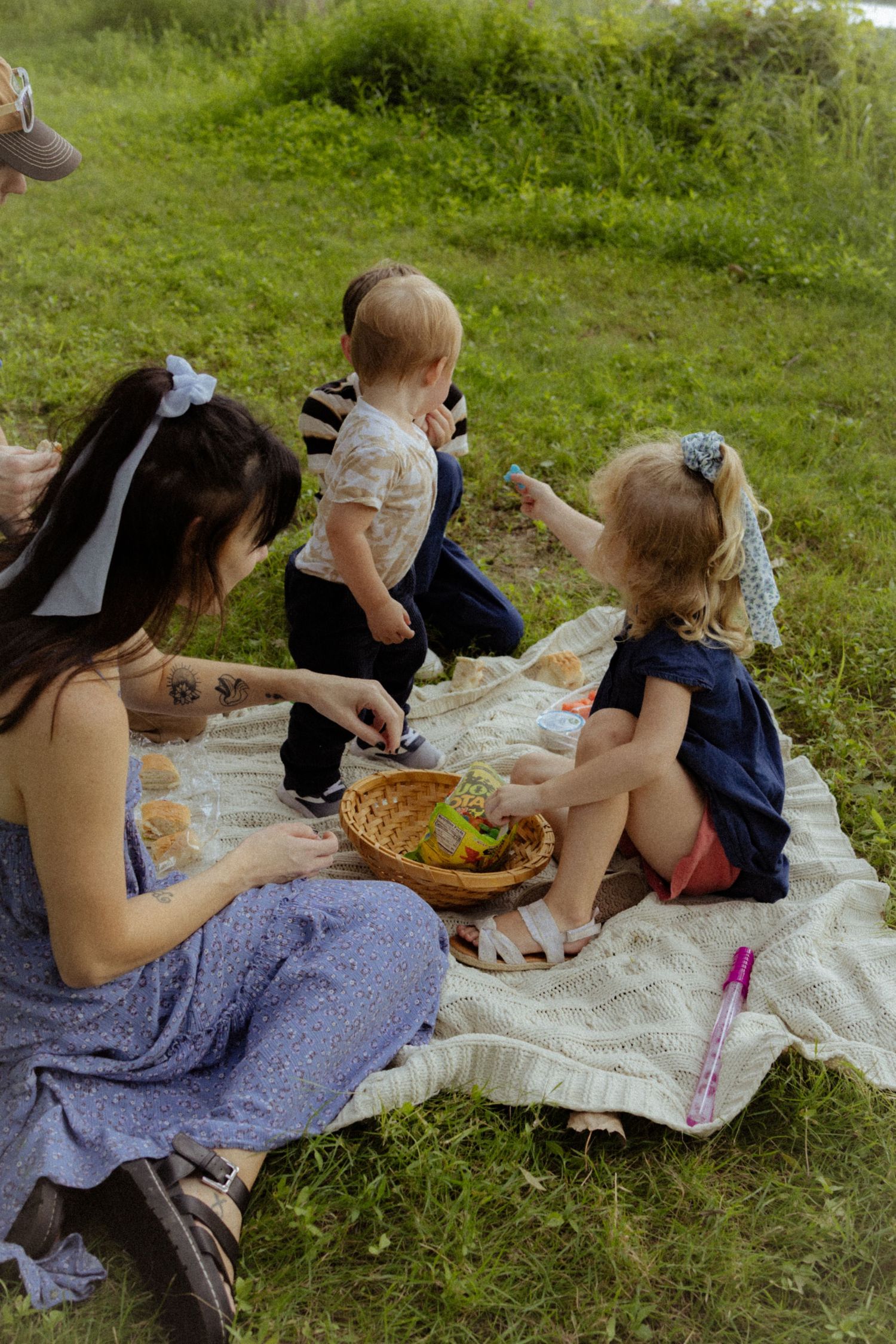 A mother and two young children enjoy a picnic on a blanket in a lush green park.