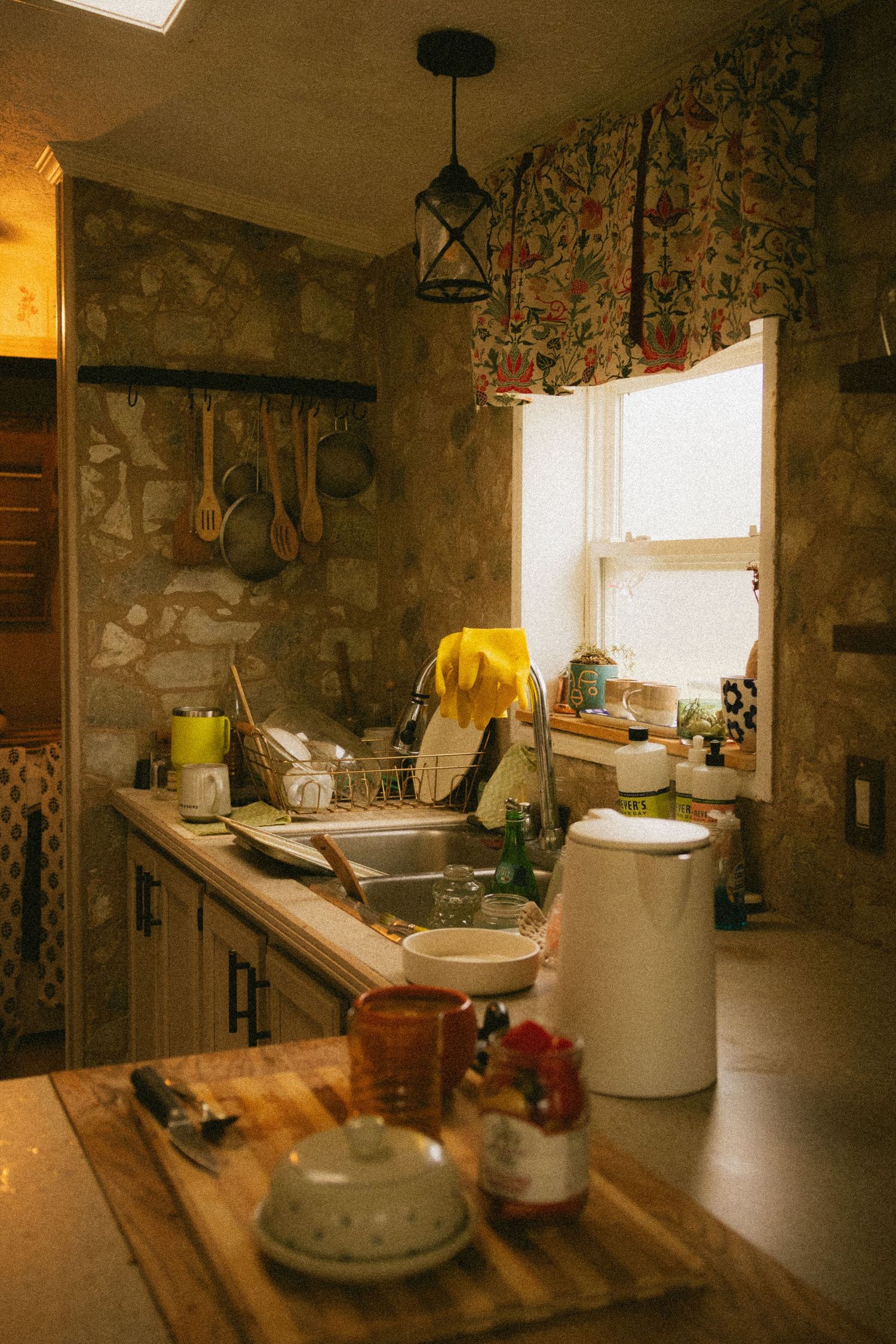 Rustic stone kitchen with wooden countertop, hanging utensils, floral curtains, and a cluttered sink area bathed in warm light.