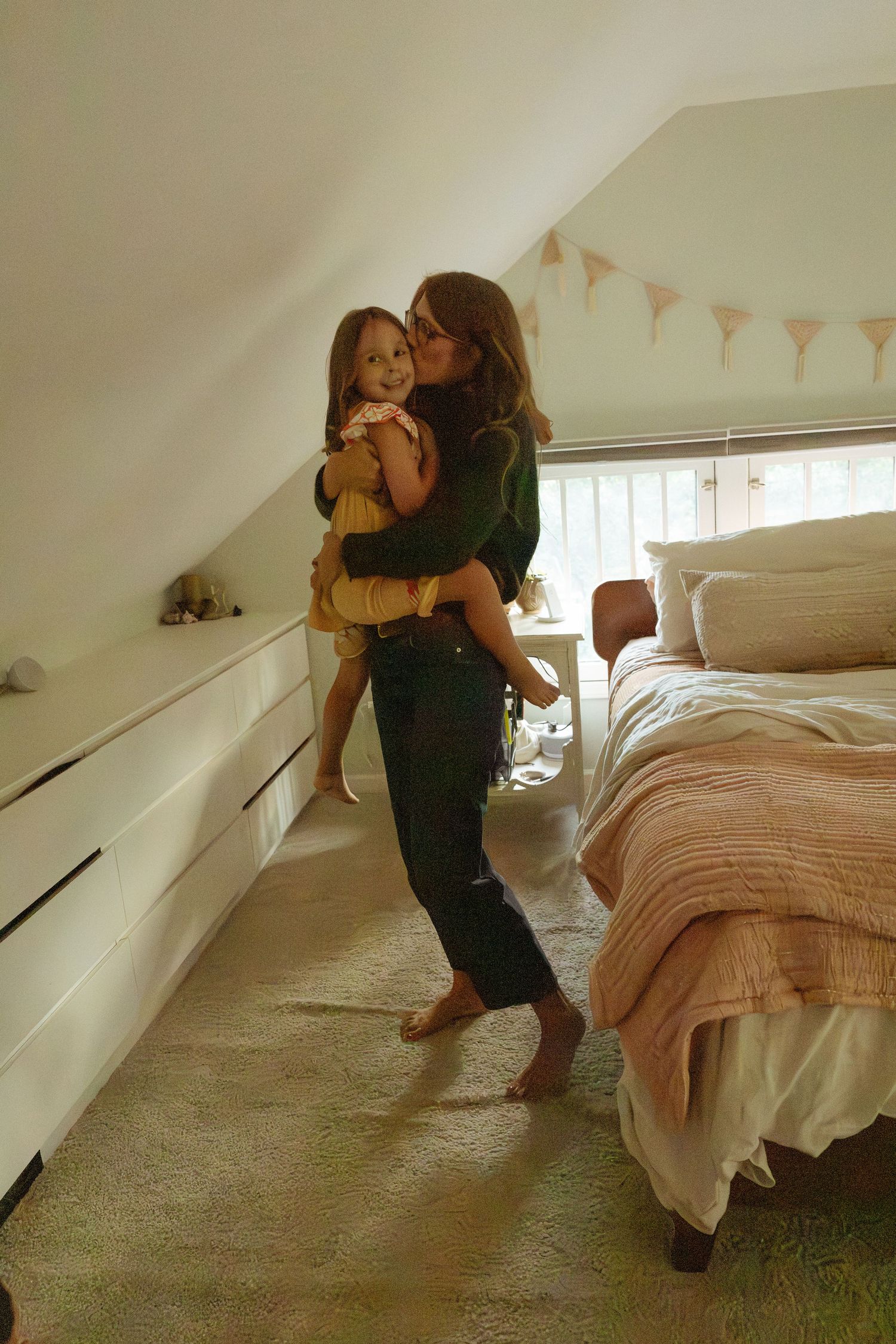 A woman in dark clothes holds a smiling young girl in a cozy attic bedroom with a sloped ceiling, pink blanket, and pastel bunting.