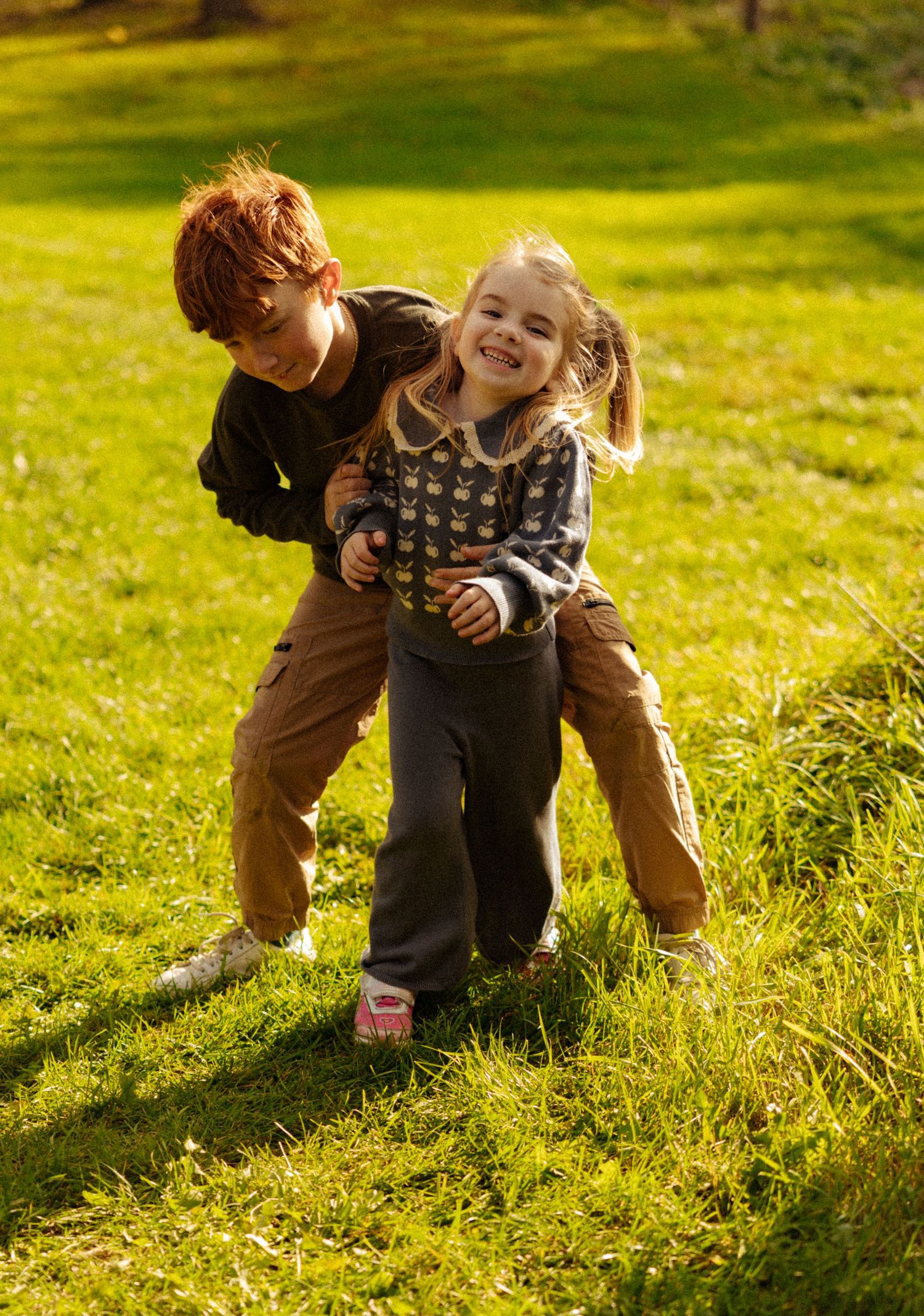 Redhead boy playfully hugging smiling blonde girl on sunny green grass outdoors.