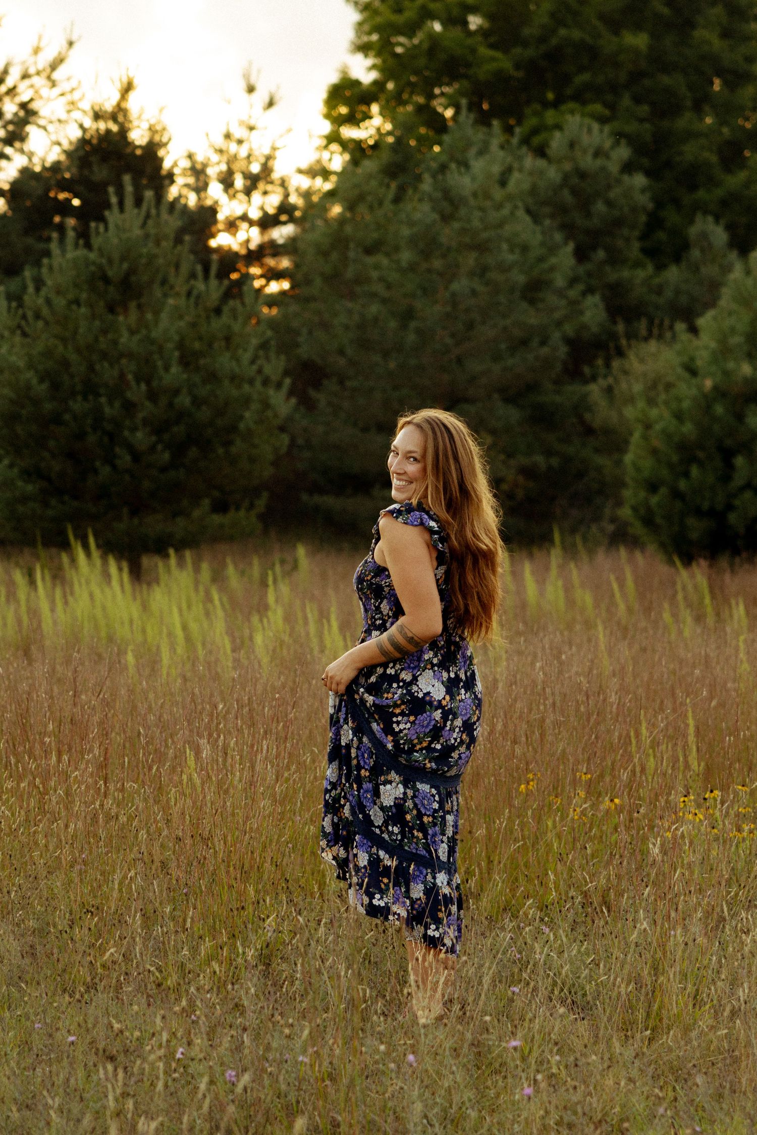 Woman in floral dress standing in golden field at sunset, smiling over shoulder with evergreen trees behind.