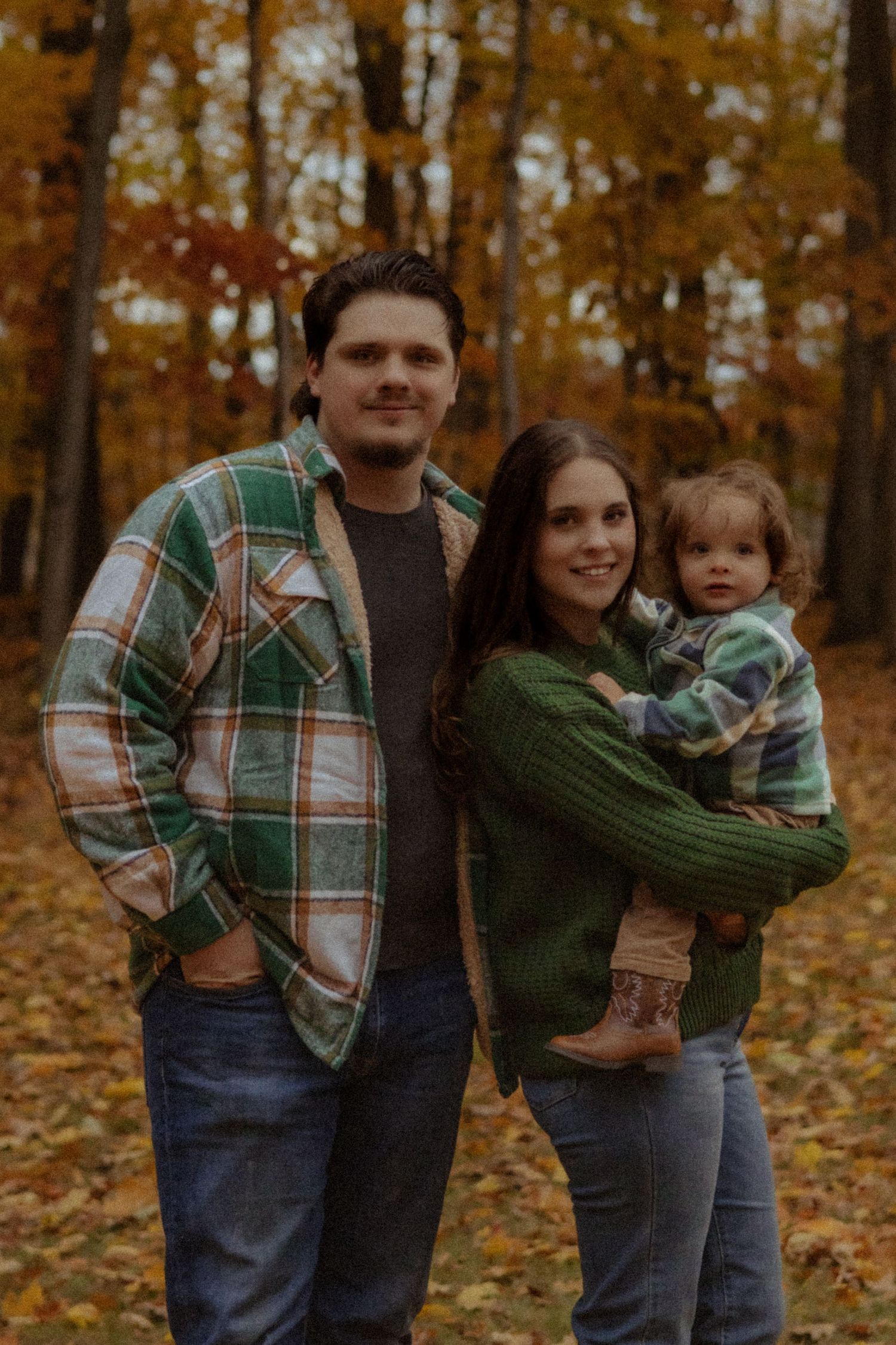 A young family of three poses in an autumn forest, wearing green and plaid flannel outfits, with colorful fall foliage behind them.