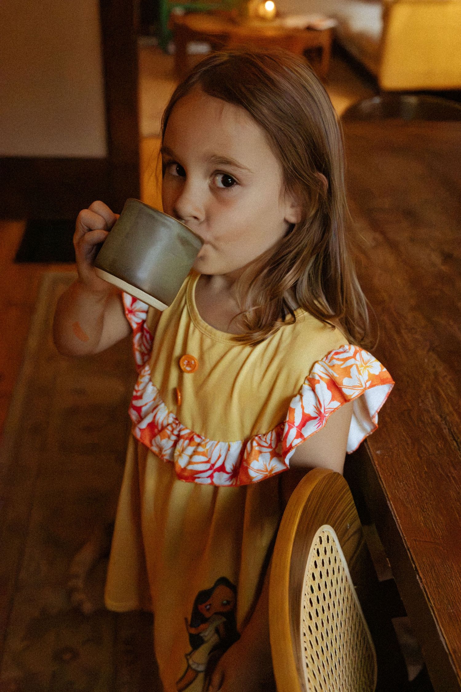 Young girl in a yellow dress with orange floral ruffle sleeves sips from a gray ceramic mug while seated at a wooden table.