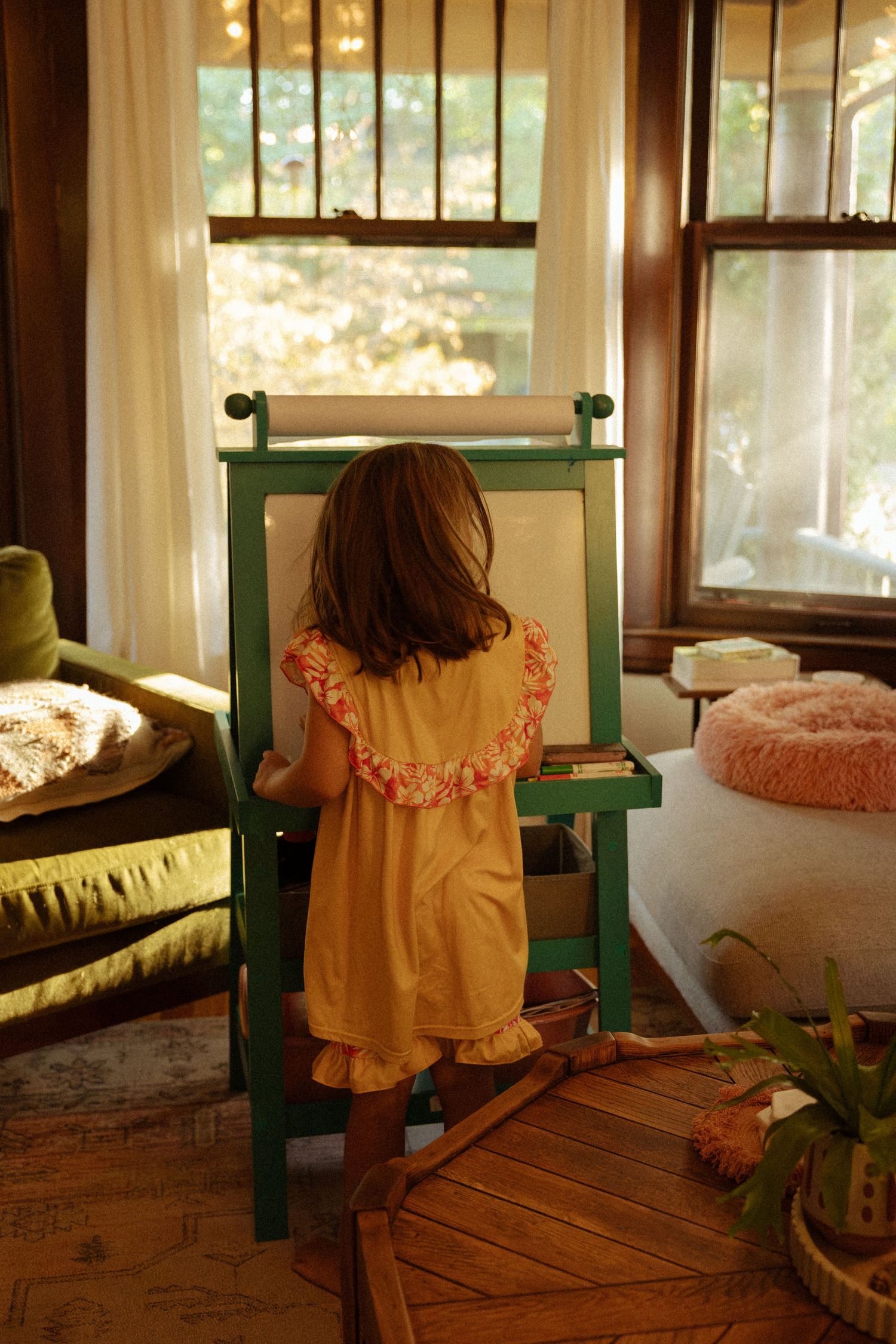 Young girl in a yellow ruffled dress stands at a green wooden art easel in a sunlit living room with large windows and cozy furniture.