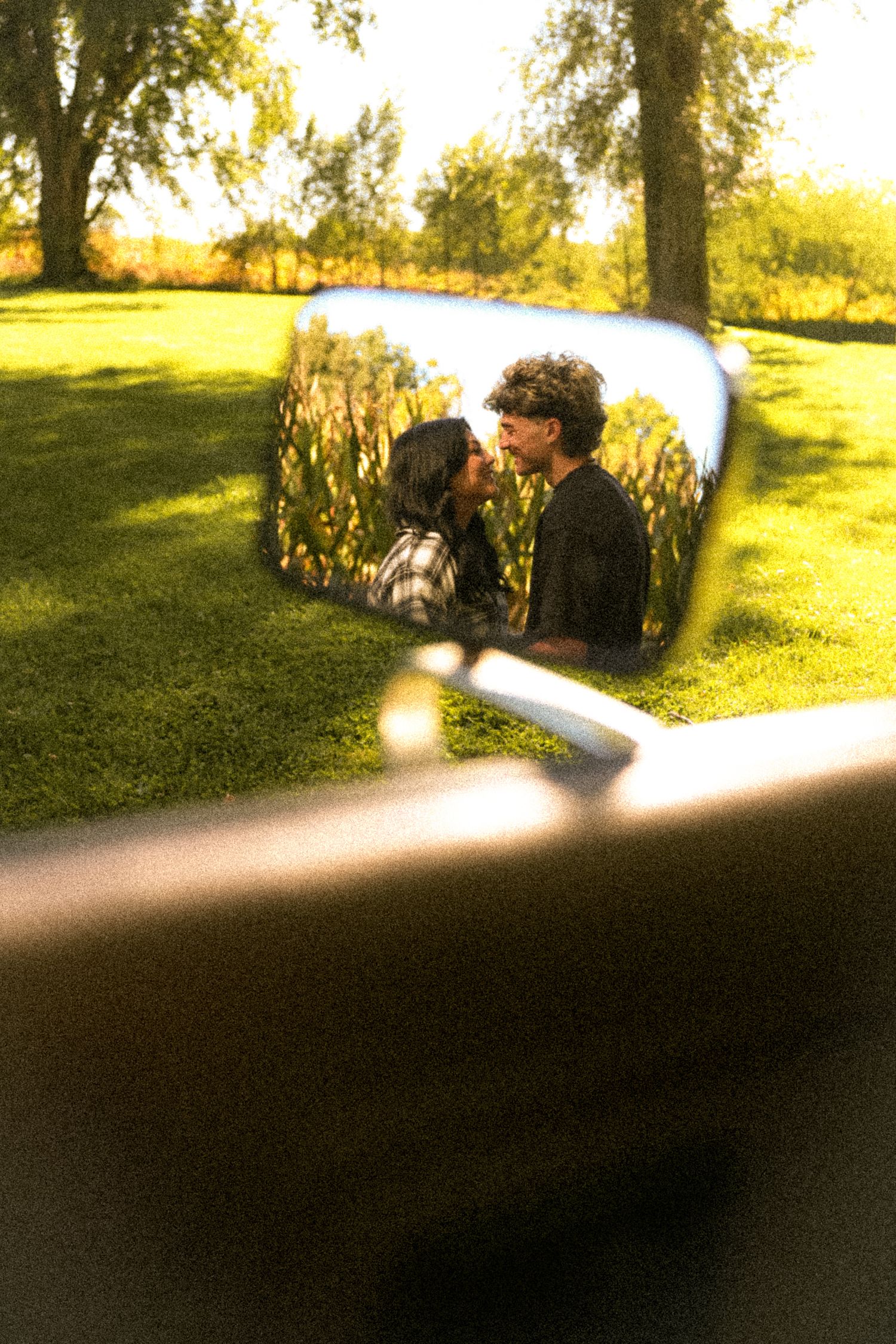 Couple sharing a romantic kiss reflected in a motorcycle side mirror, surrounded by lush green park trees.