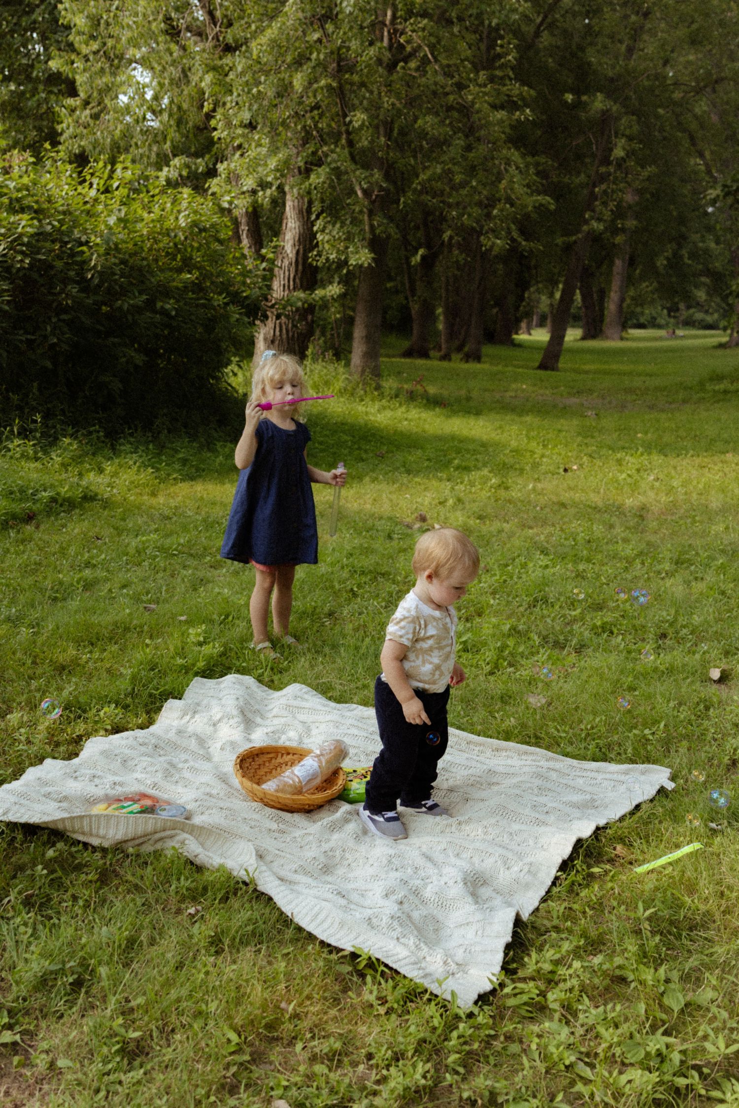 Two young siblings playing on a white blanket during a picnic on green grass in a shaded park.