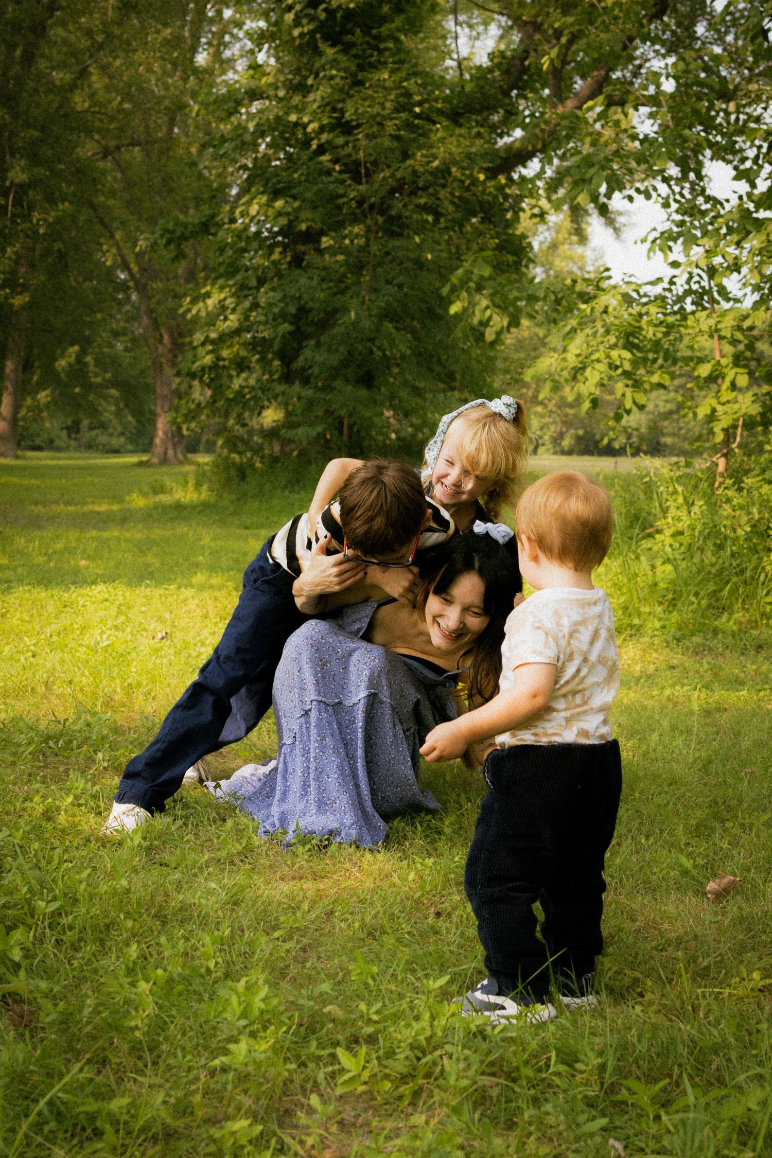 A mother kneels on green grass in a park, joyfully hugging her three young children on a sunny day.