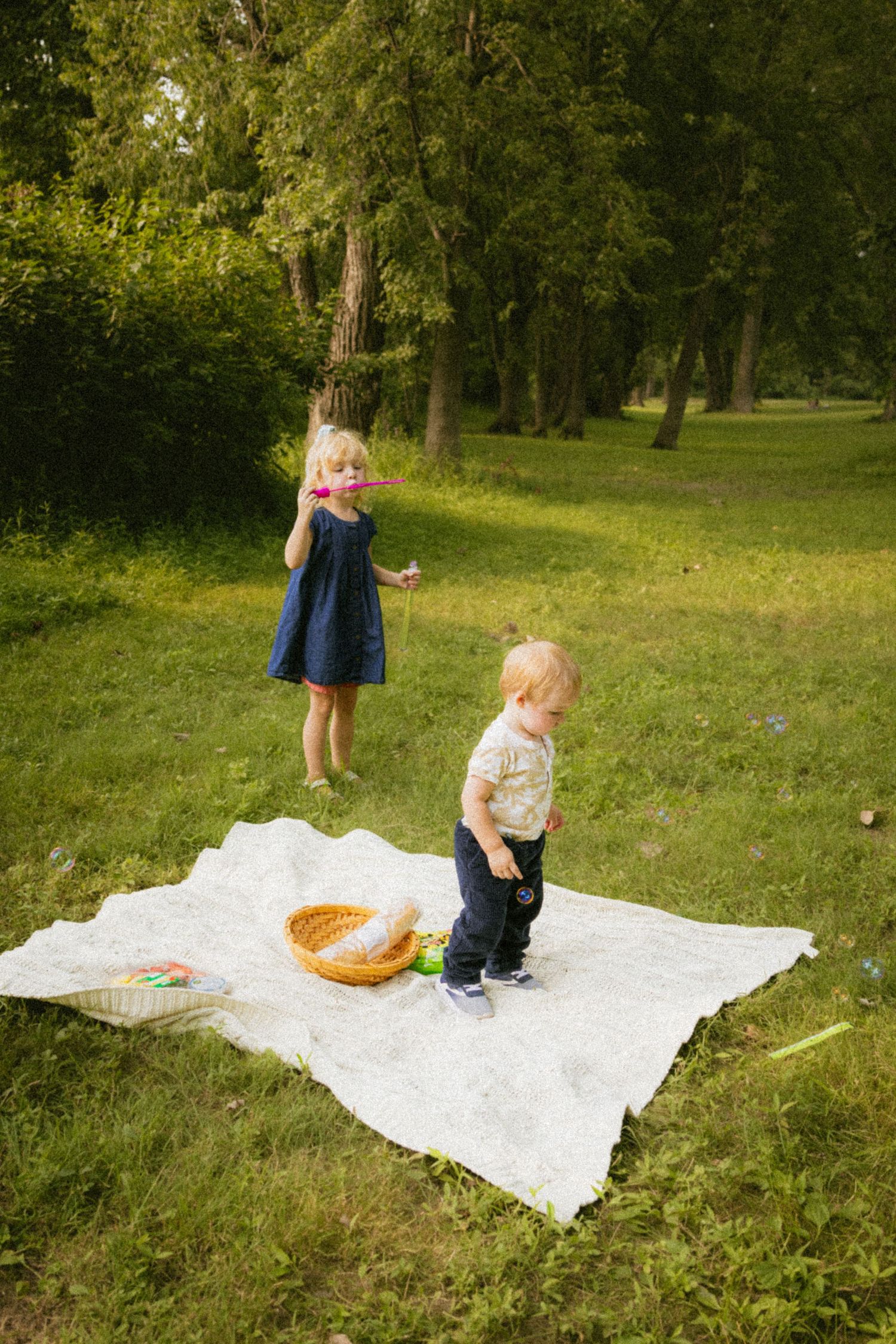 Two young children playing on a white blanket during a sunny outdoor picnic in a lush green park.