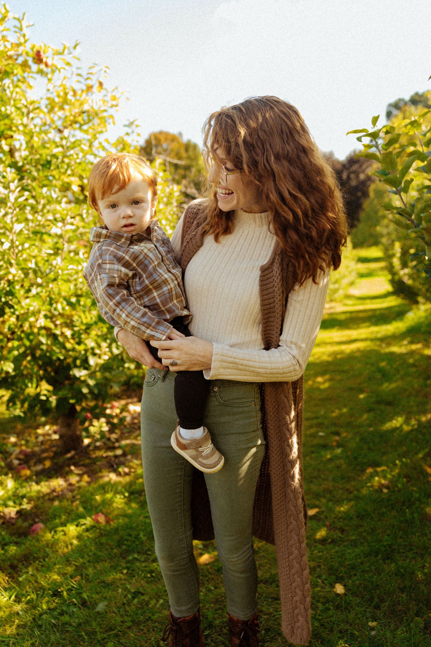 A smiling mother with curly hair holds her red-haired toddler in a sunny apple orchard during fall.