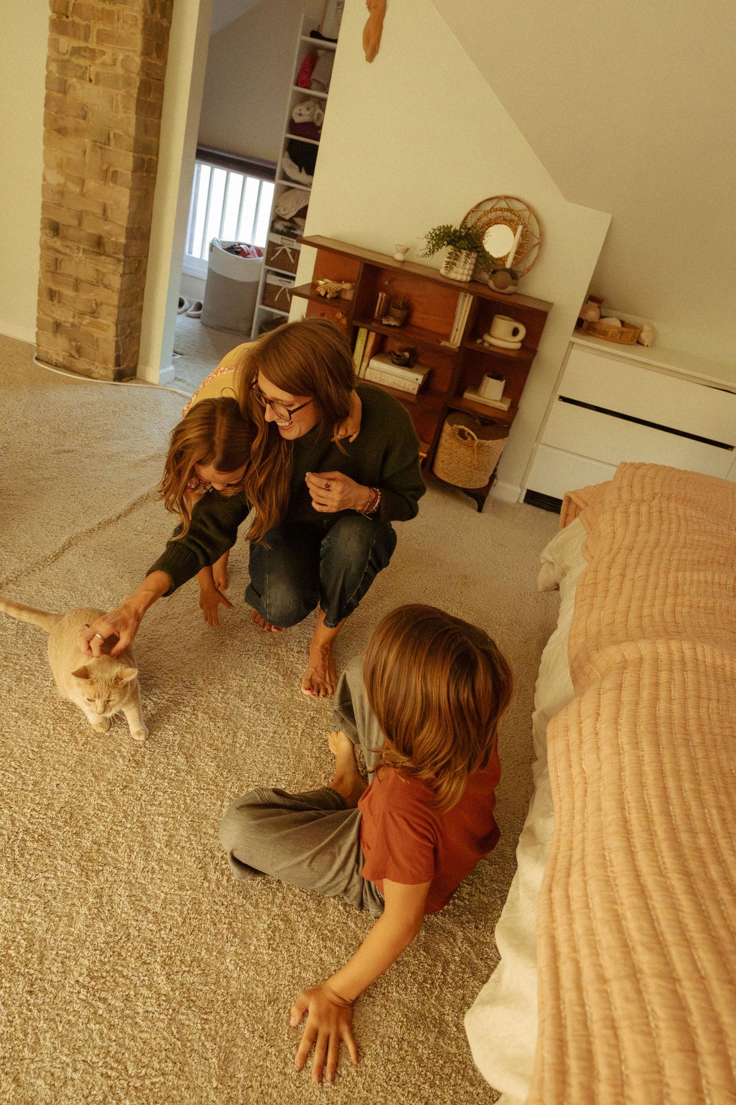 A woman and two children sit on a cream carpet in a cozy attic bedroom, gently petting a small orange cat near a bed.