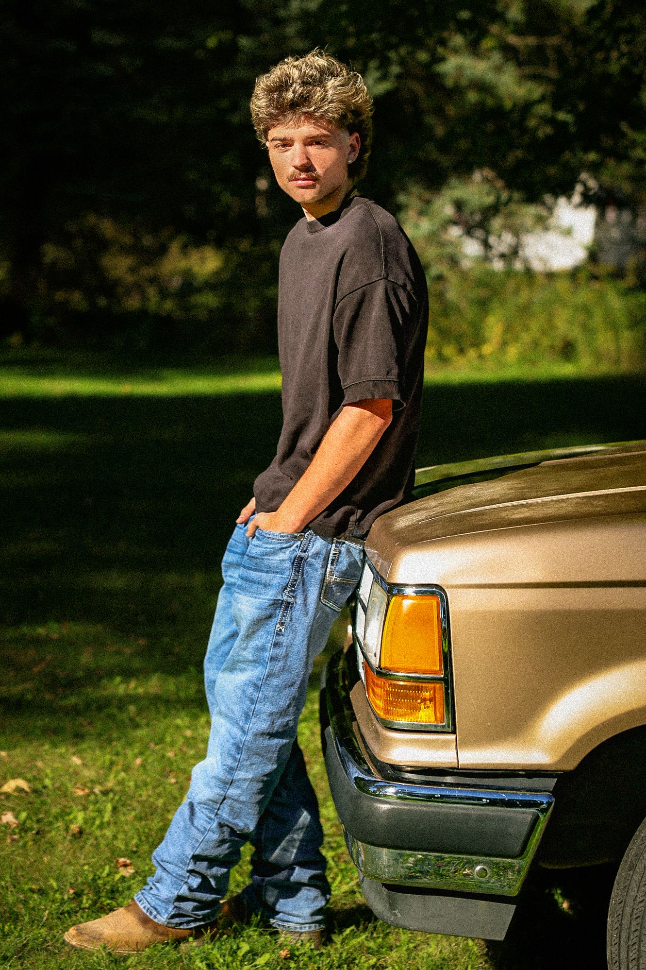 A teenage boy in a black t-shirt and jeans leans against a gold truck on a sunny day with green trees behind him.
