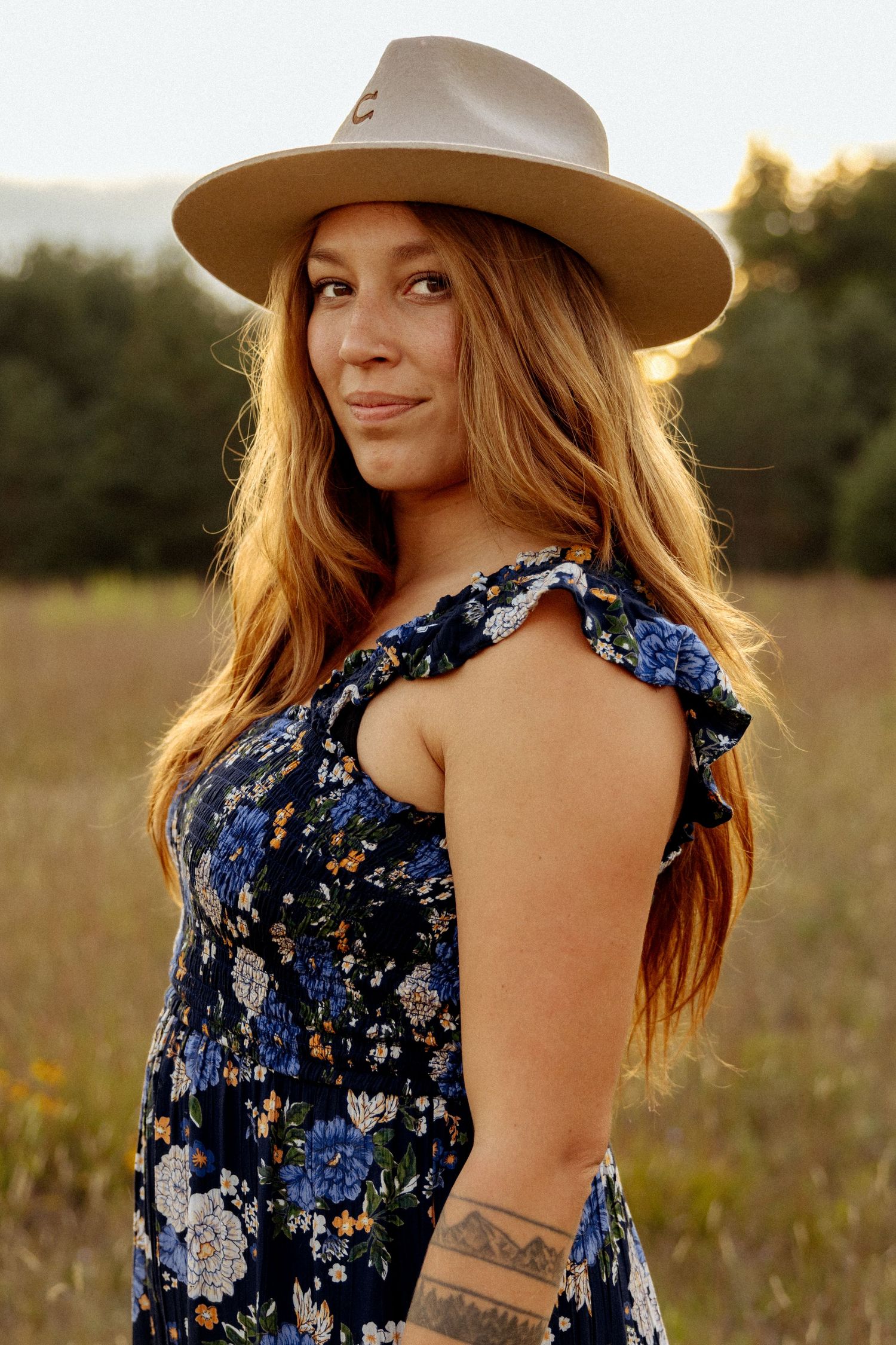 Woman in floral dress and tan wide-brim hat posing in golden hour field with long auburn hair.