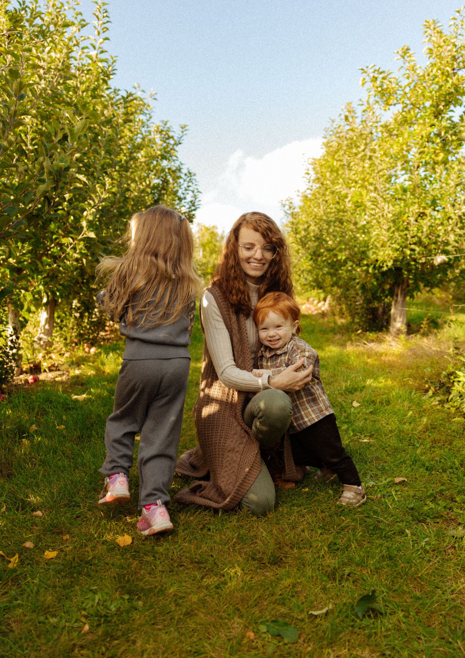 Smiling mother kneeling in apple orchard hugging red-haired toddler while young girl stands beside them.
