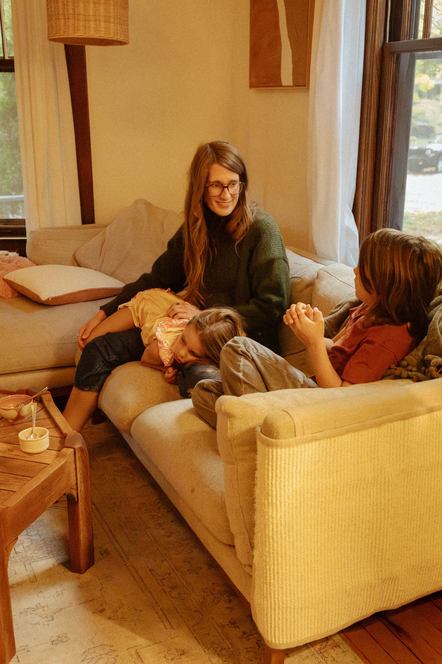 A woman with glasses relaxes on a cozy cream sofa with two children in a warmly lit living room with a wooden coffee table.